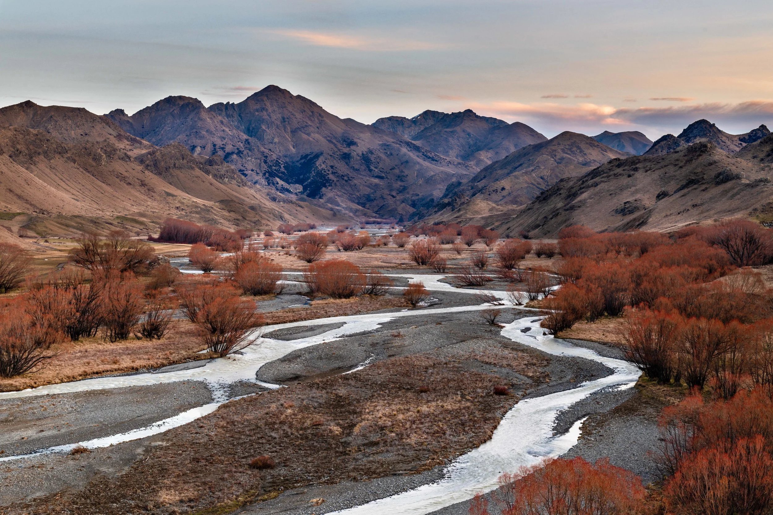 A panoramic view of a mountain valley with a winding river, sparse orange and brown bushes, and distant rugged mountains under a partly cloudy sky.