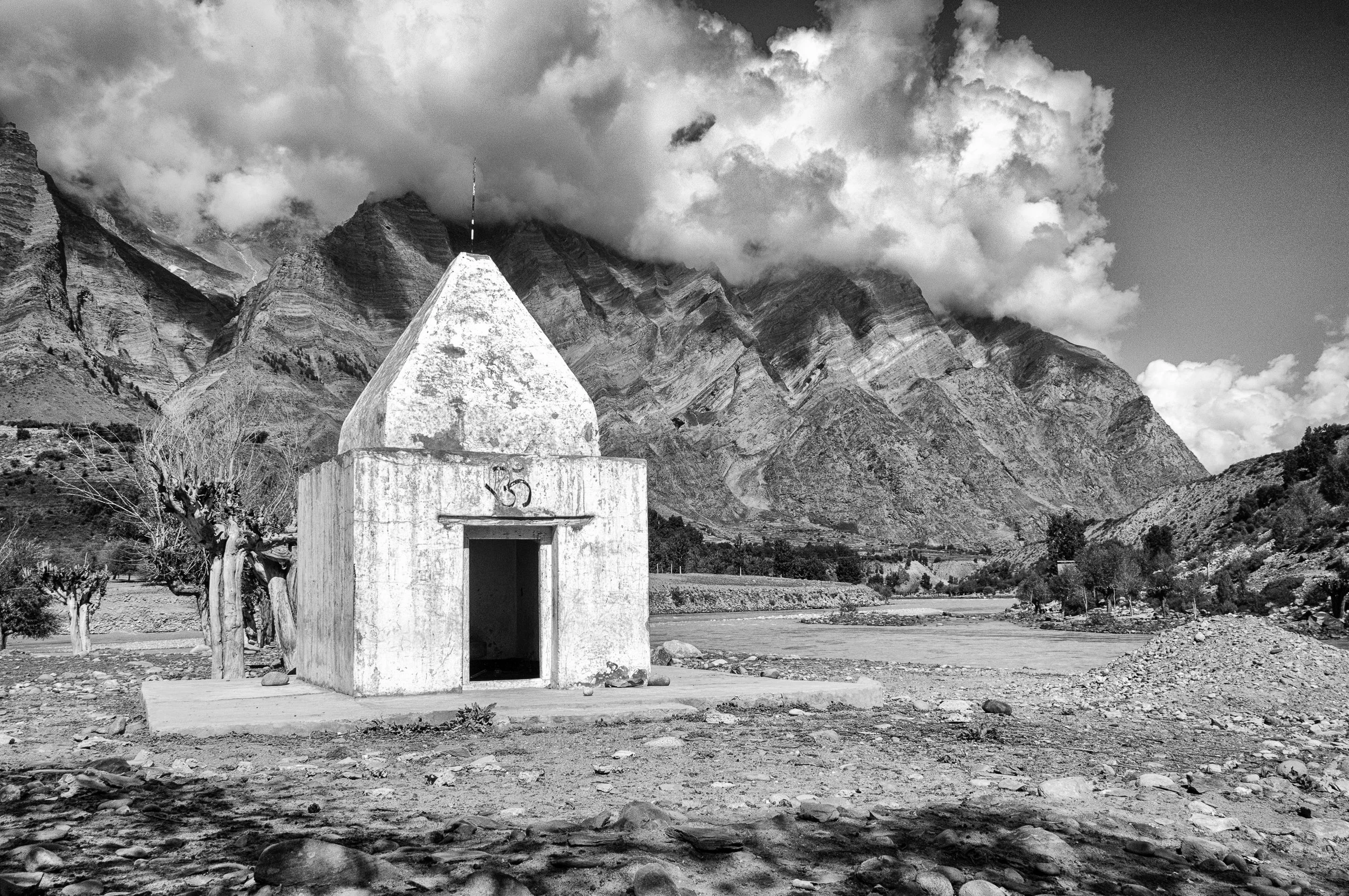 Small white chapel with a pointed roof and an open door, set against a mountainous landscape with clouds in the sky and leafless trees nearby, in black and white.
