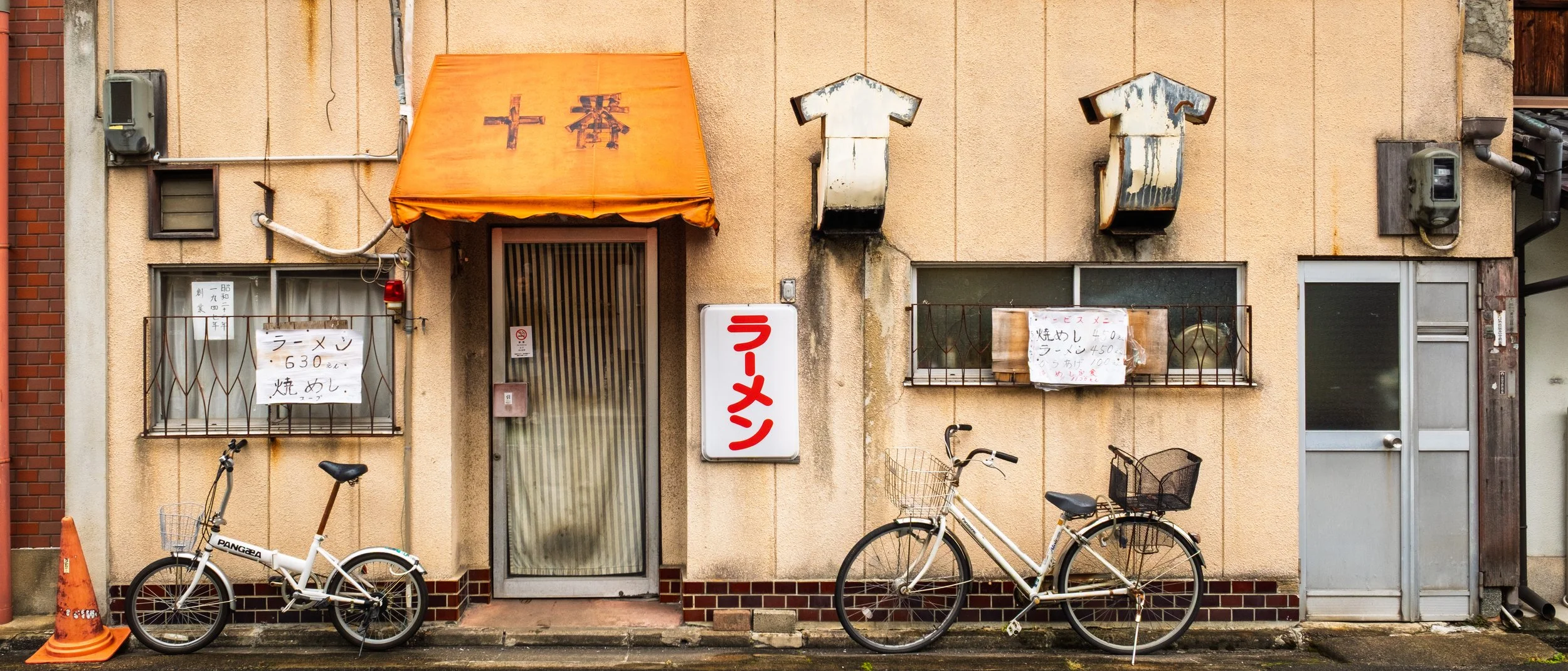 A small Japanese ramen restaurant with two bicycles parked in front, an orange awning with Japanese characters, and handwritten signs in Japanese on the windows and wall.