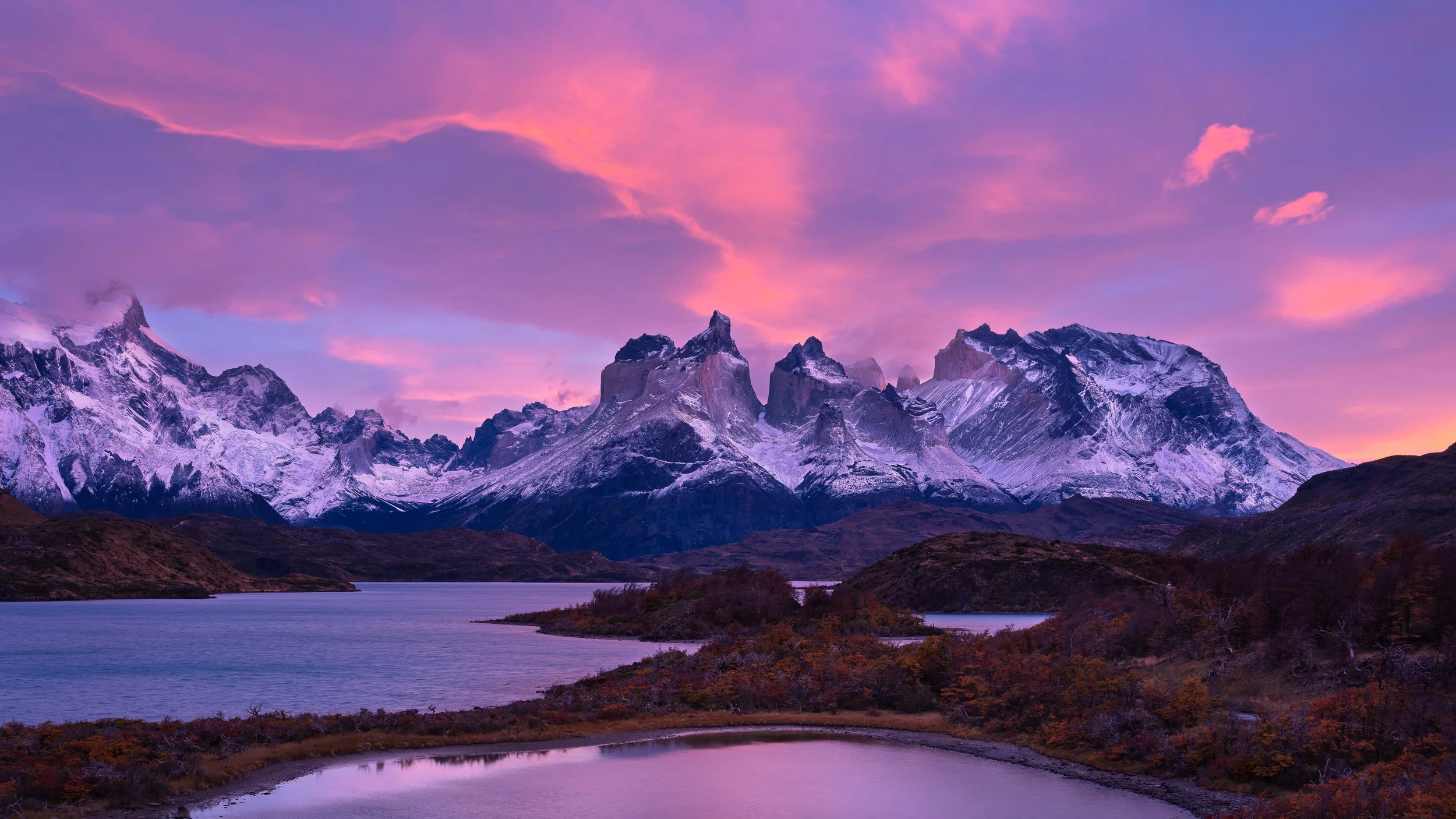 Snow-capped mountains under a colorful pink and purple sky with a lake and autumn foliage in the foreground.