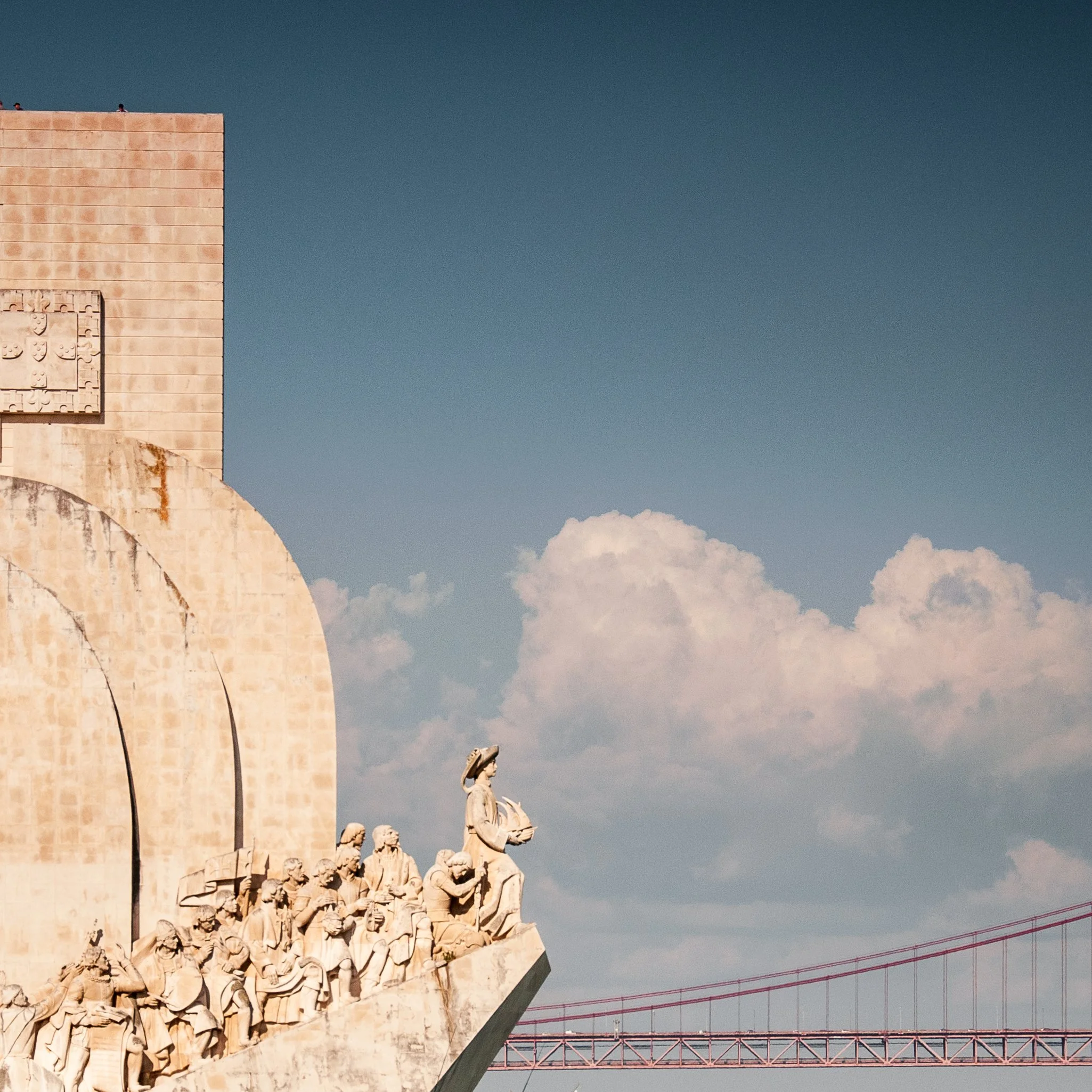 Stone monument with carved sculptures of people, a woman holding a bowl, under a blue sky with scattered clouds, and a red bridge in the background.