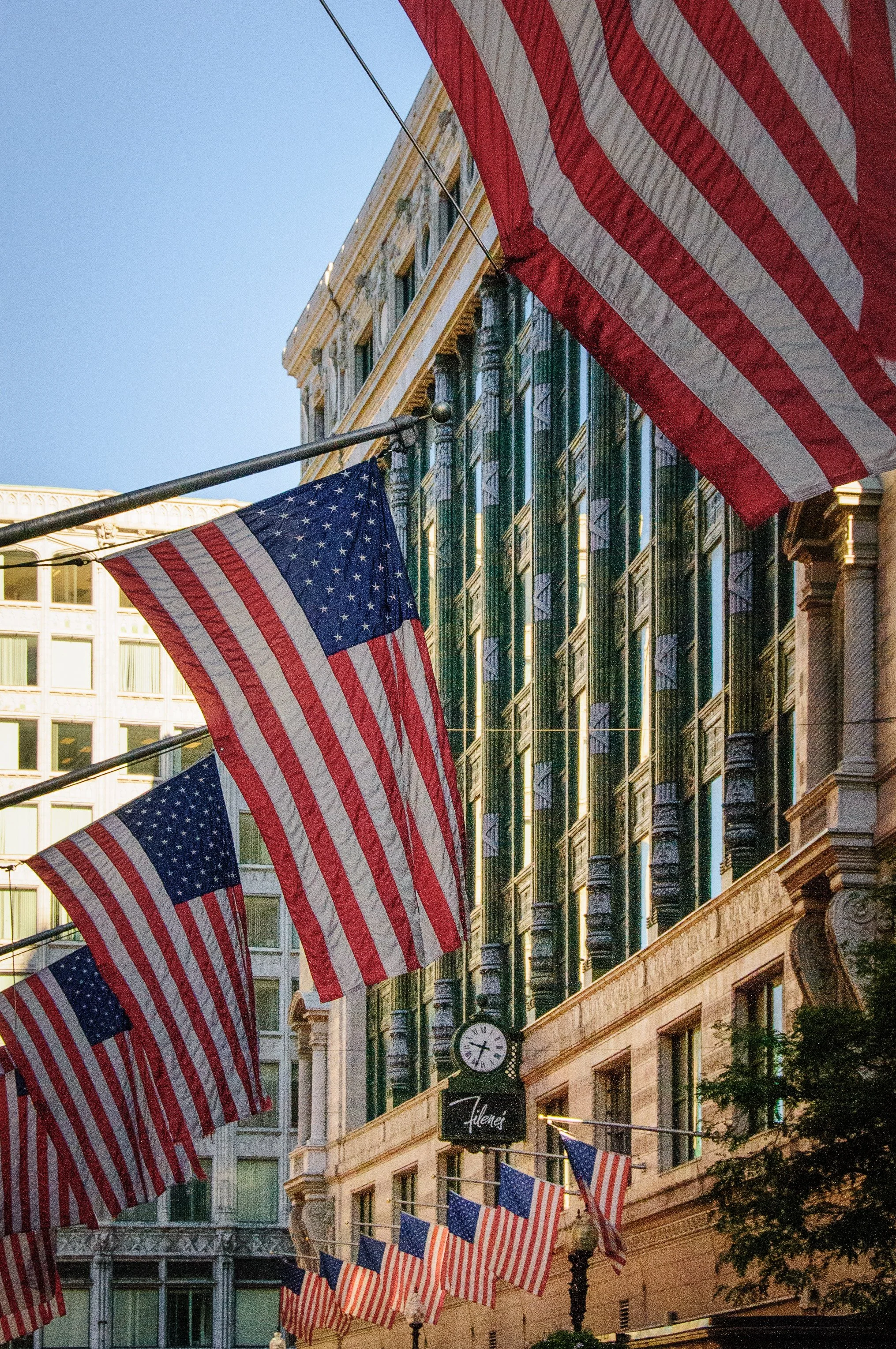 Multiple American flags hanging above a city street with historic buildings and a clock showing 8:15.