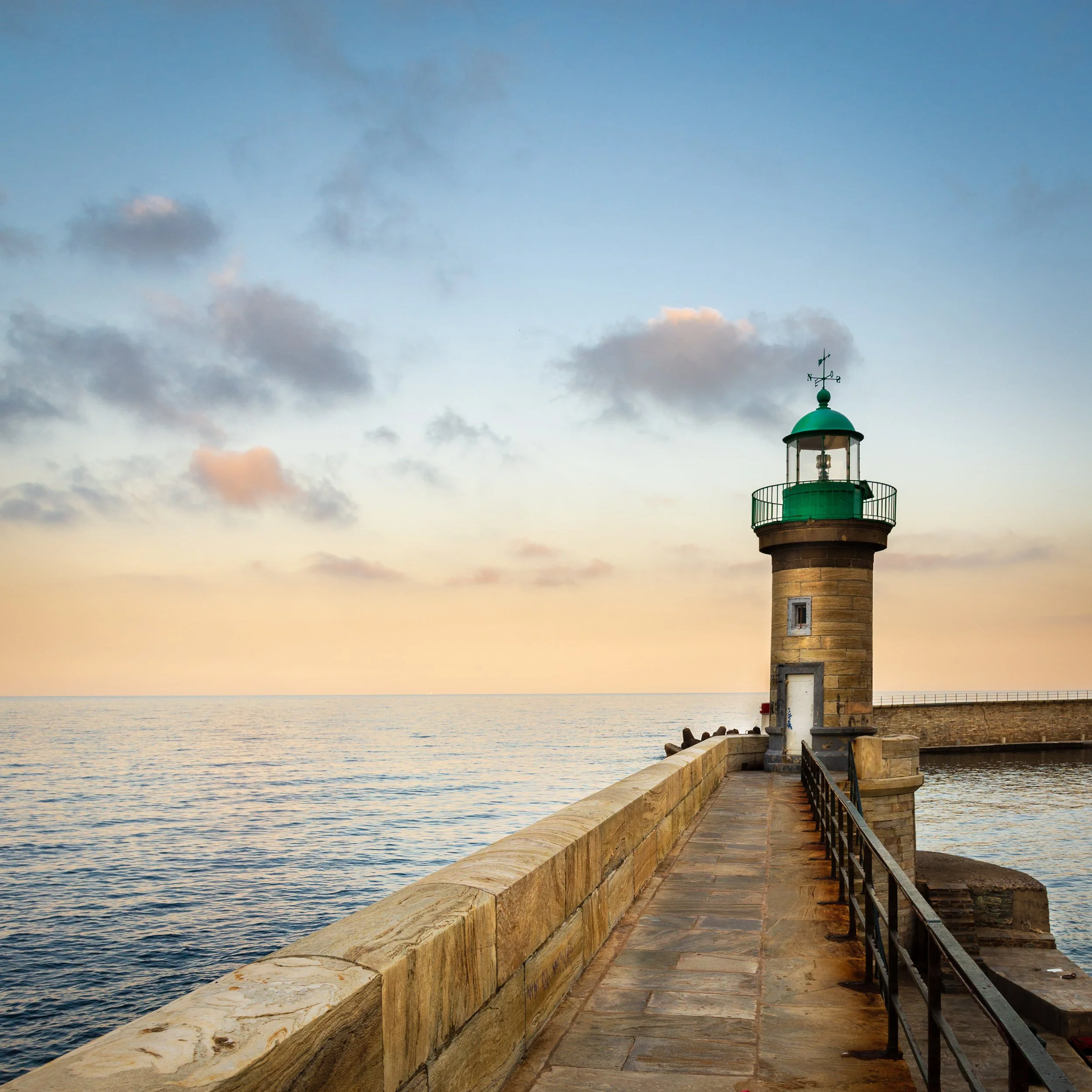 A stone lighthouse at the end of a pier overlooking calm sea during sunset with a partly cloudy sky.