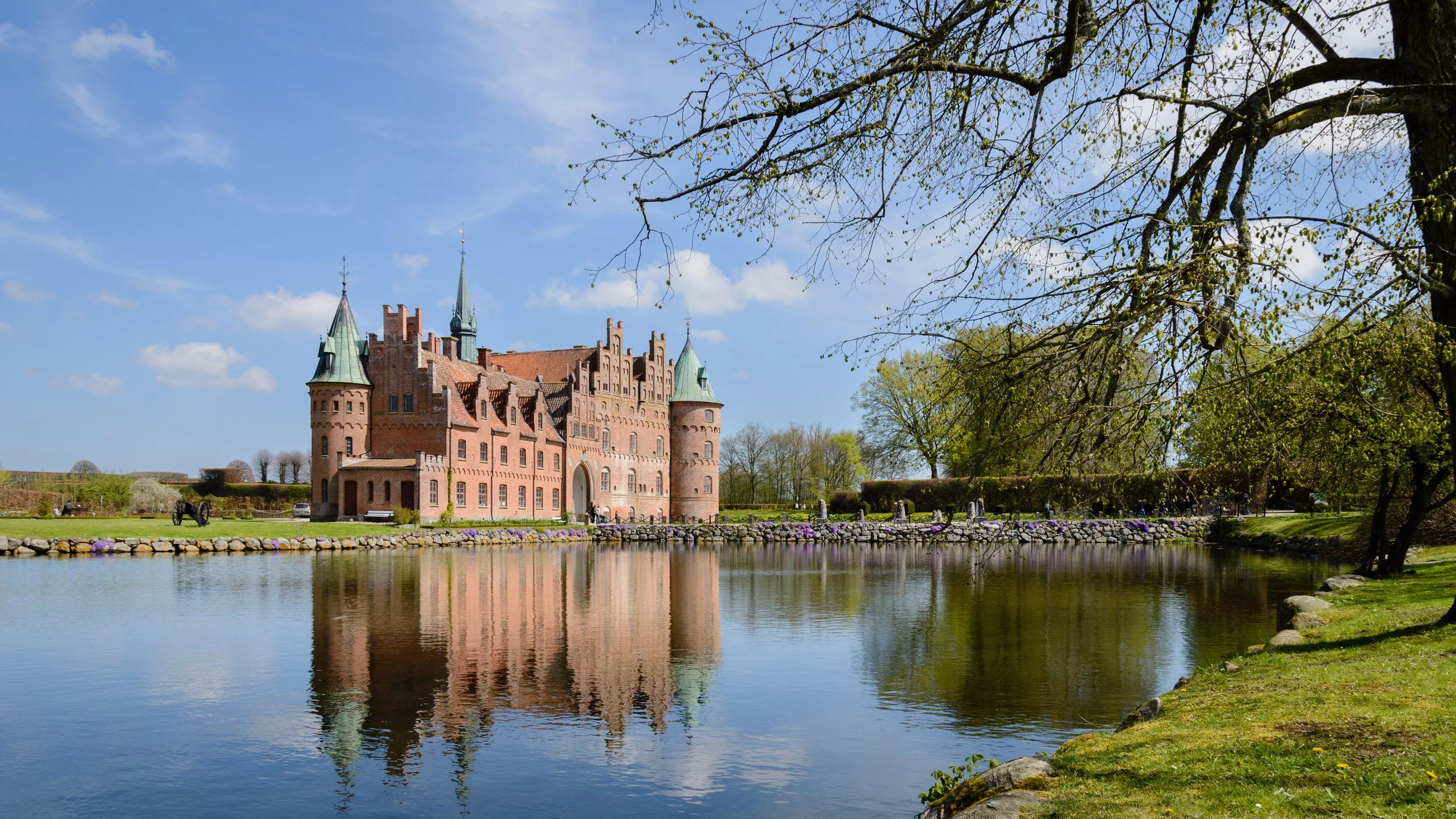 A historic castle with conical towers and brick walls, situated by a calm river with a stone border. The sky is blue with some clouds, and trees with budding leaves frame the scene.