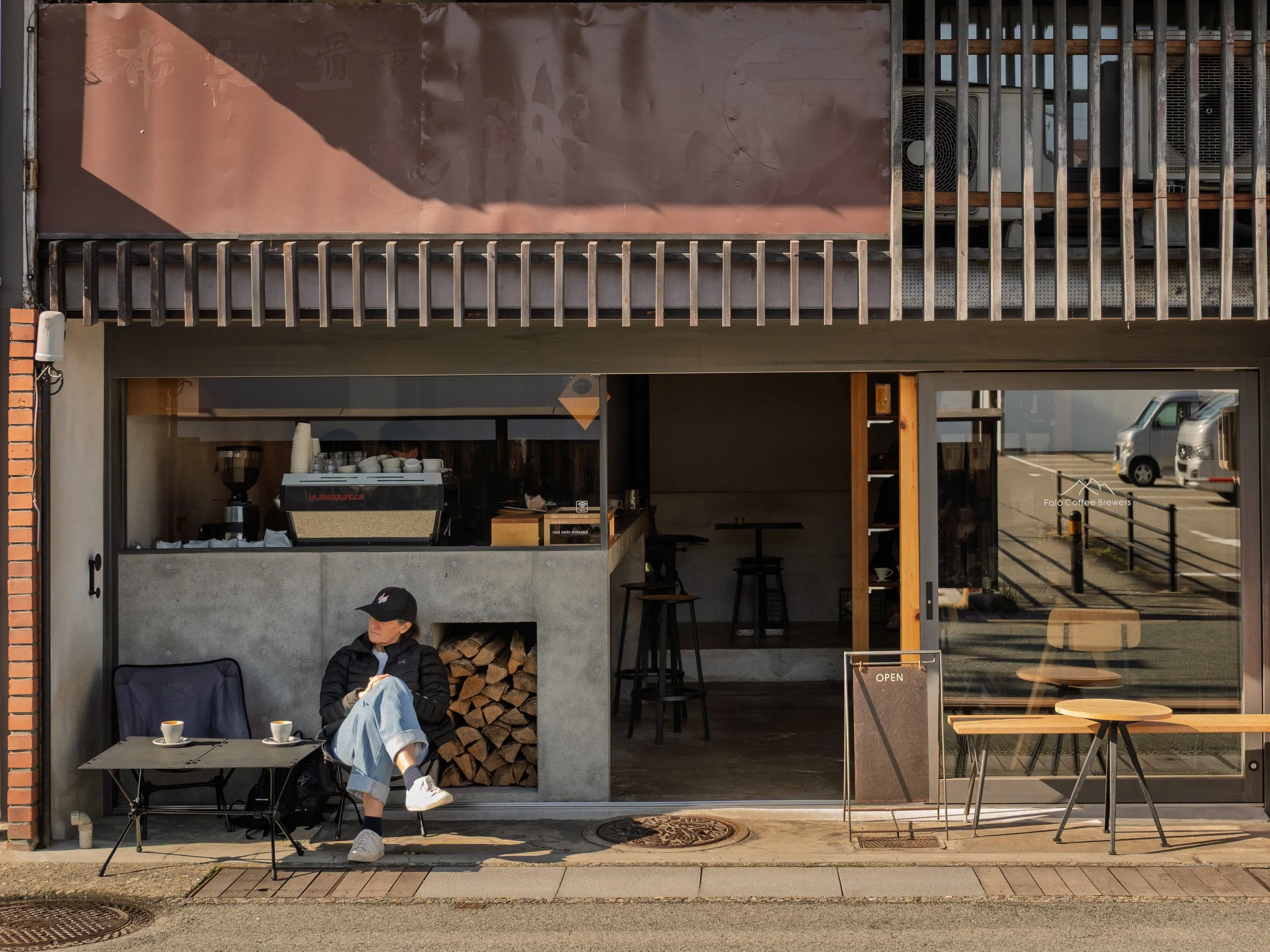 A person is sitting outside a modern coffee shop on a small chair, drinking coffee. The shop has a glass front showing a coffee machine inside, and a sign indicating it is open. There is a stack of firewood and some tables and chairs outside.