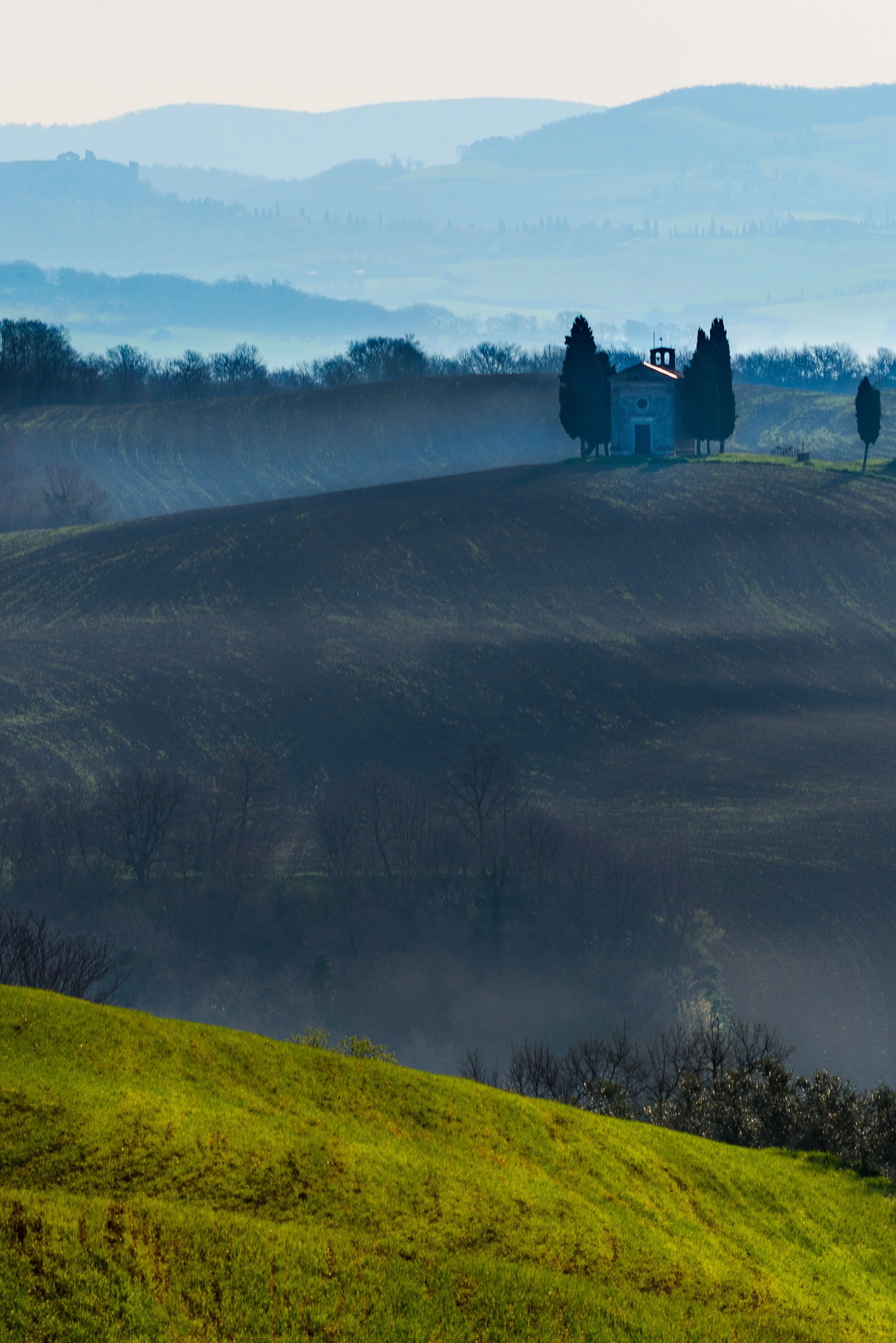 A small stone chapel surrounded by tall cypress trees on a hilltop in the countryside, with rolling hills and layers of misty landscape in the background.