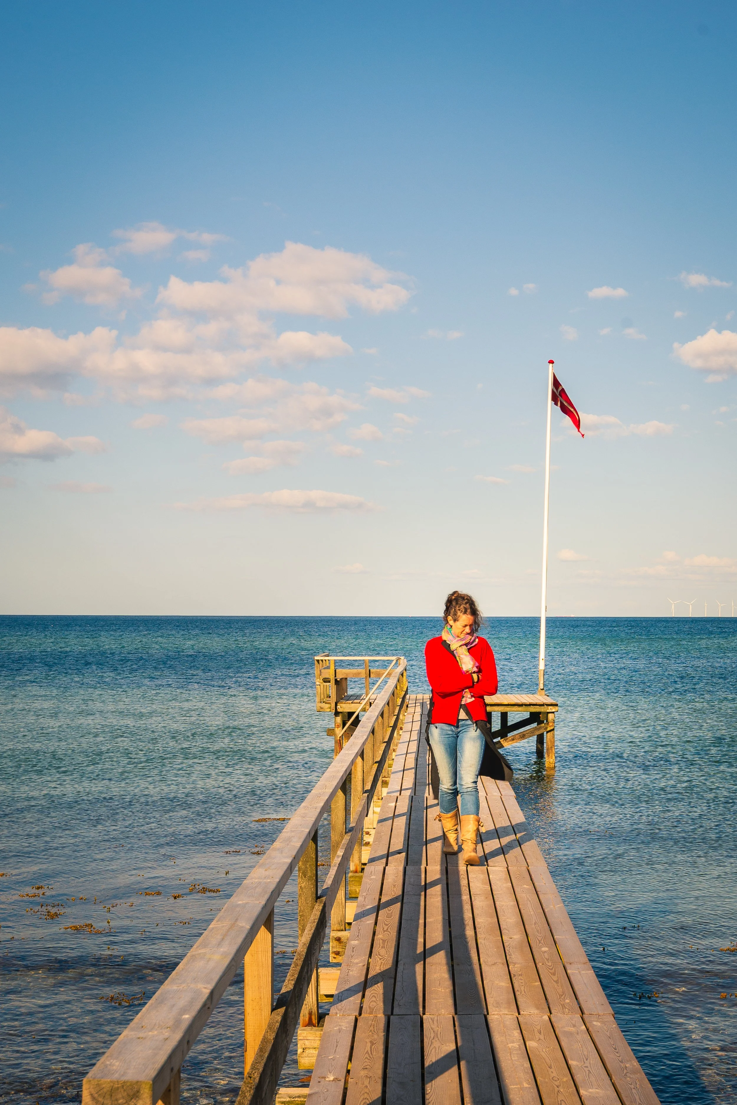 A woman walking on a wooden pier over the ocean, wearing a red coat and tan boots, with a flag waving on a pole nearby, under a partly cloudy sky.