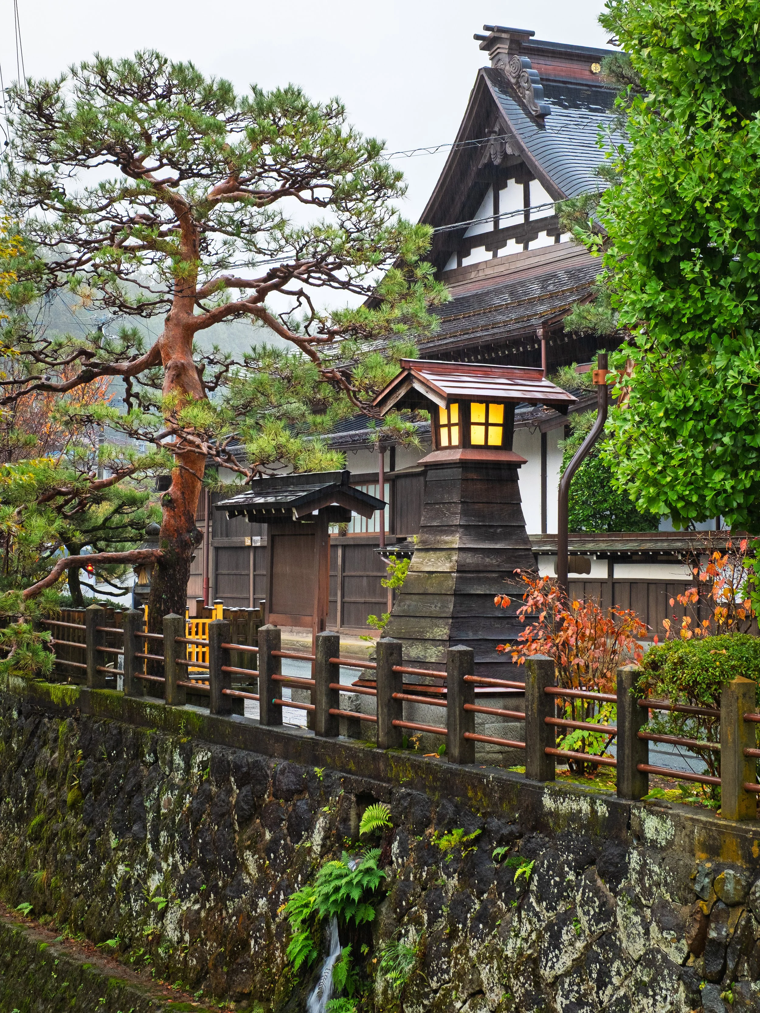 Traditional Japanese building with a tiled roof, wooden fence, a stone wall with moss, and greenery including a pine tree and bushes.