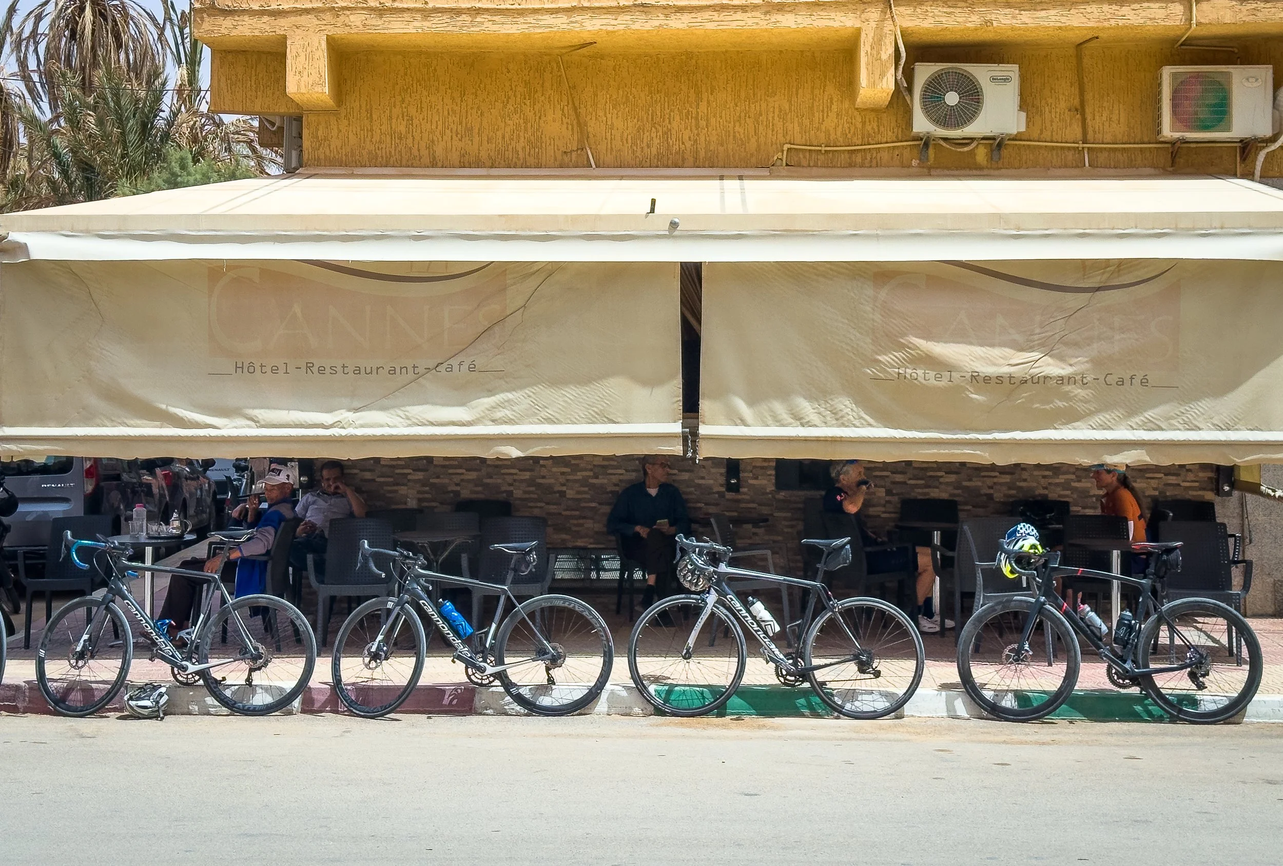 Bicycles parked outside a restaurant with people sitting at tables under a beige canopy, with a hotel sign that says 'CANNES'.