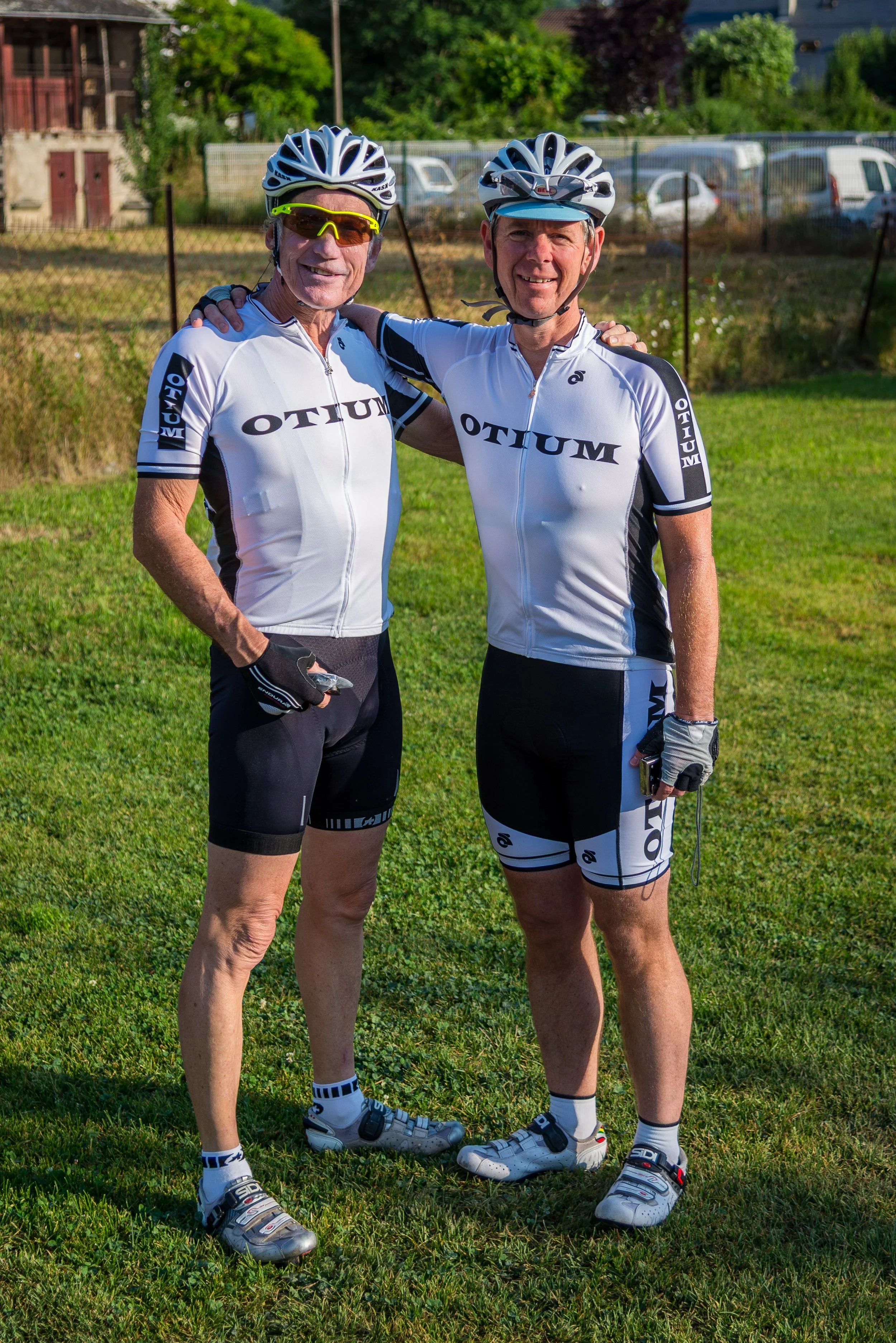 Two older men in cycling gear standing on grass with trees, a fence, and parked cars in the background. They are smiling, with one man's arm around the other's shoulder, both wearing white and black cycling jerseys, helmets, and sunglasses.