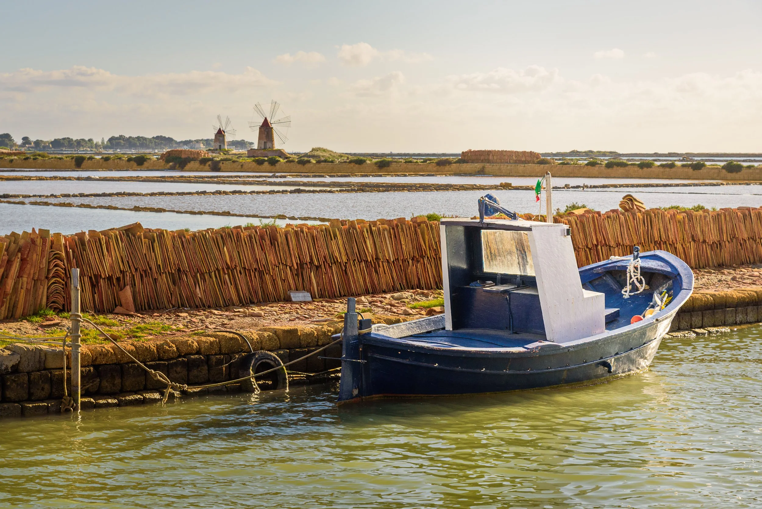 A small blue boat moored beside a stone pier, with a terracotta tiled barrier and saltwater marshes in the background, along with windmills and a partly cloudy sky.