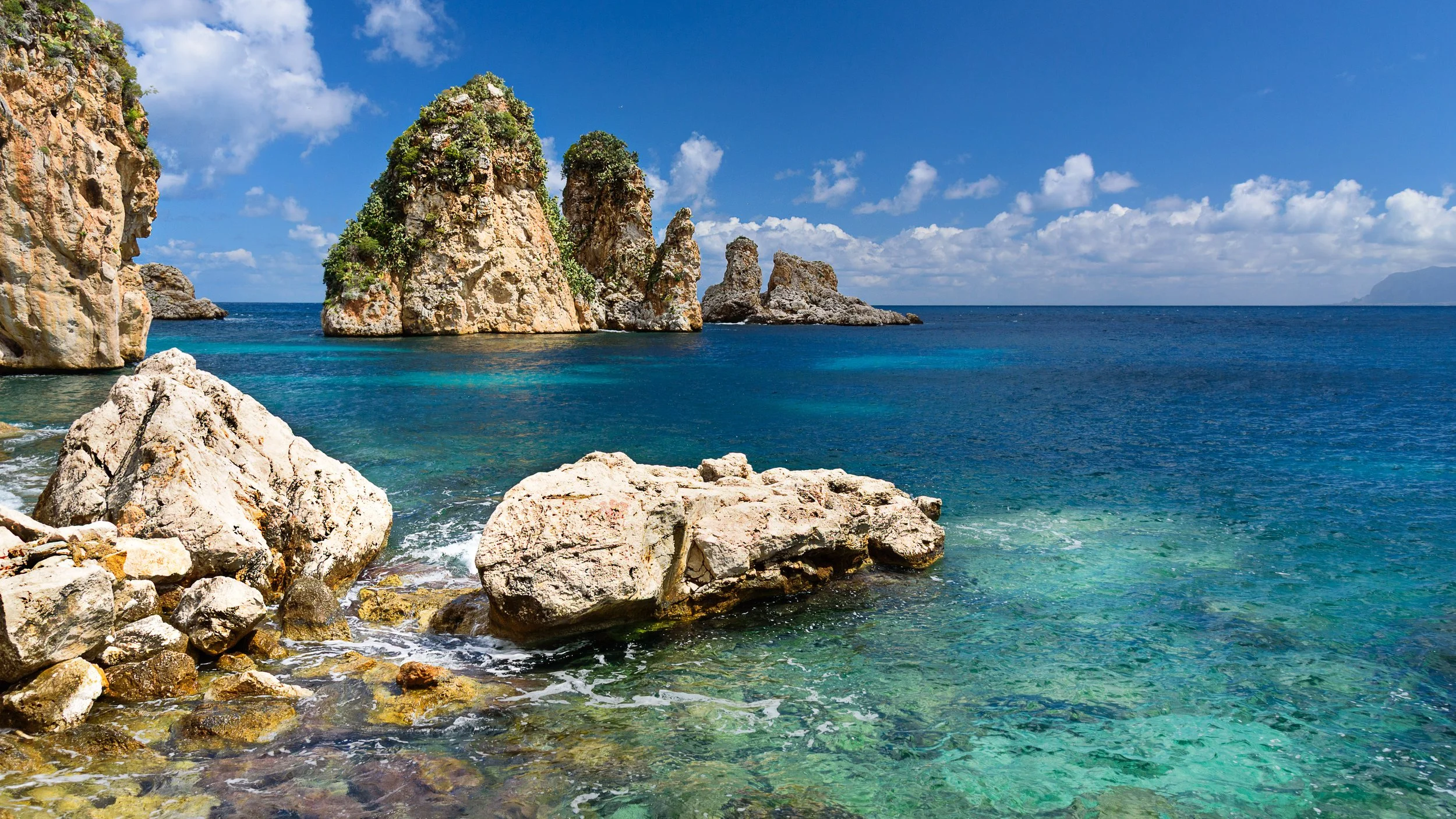 Clear turquoise sea with rocks and large rocky islands under a partly cloudy sky.