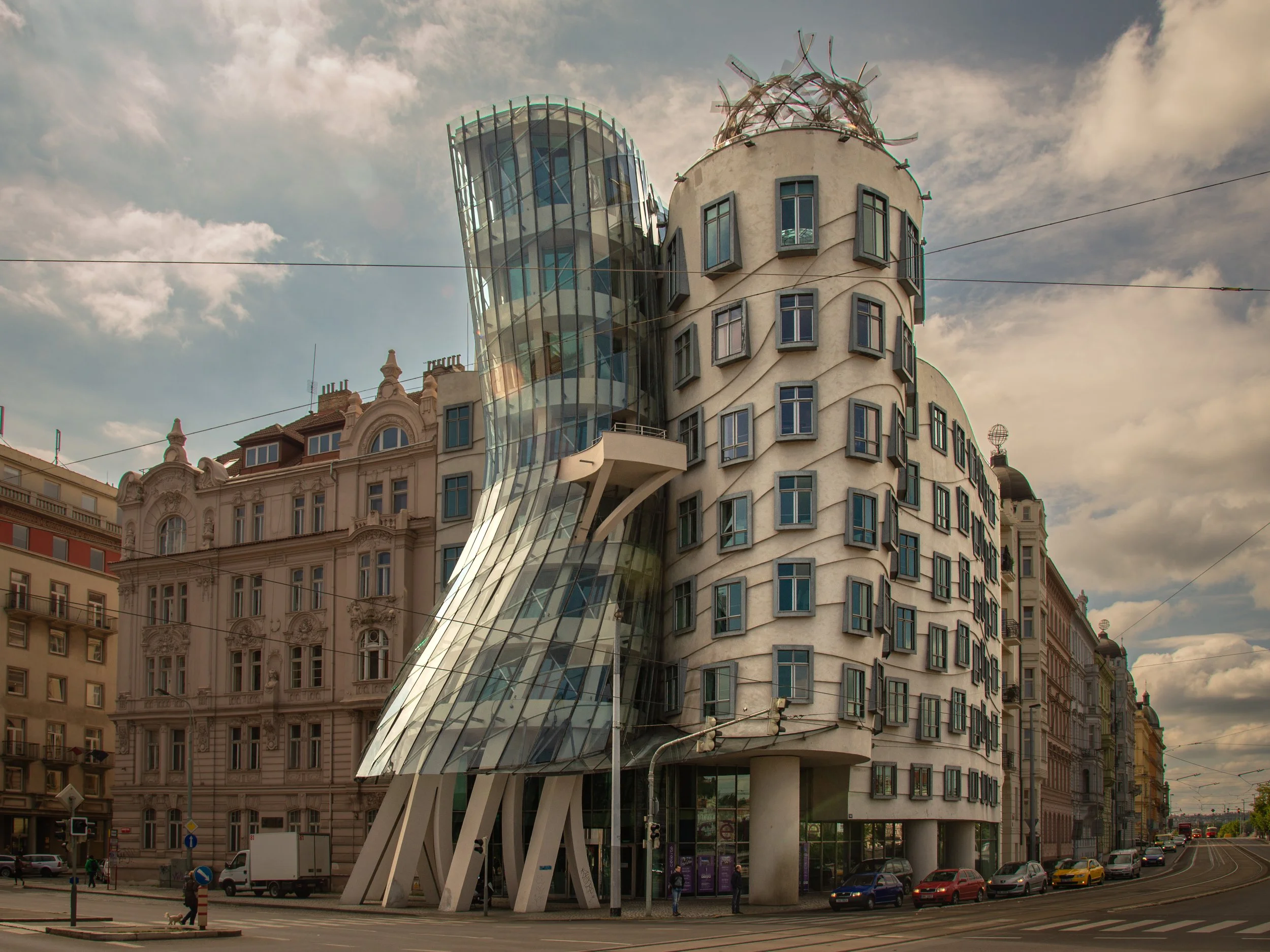 Modern building with curved glass and irregularly arranged windows, resembling a sculpture, located on a city street with parked cars and historic buildings in the background.