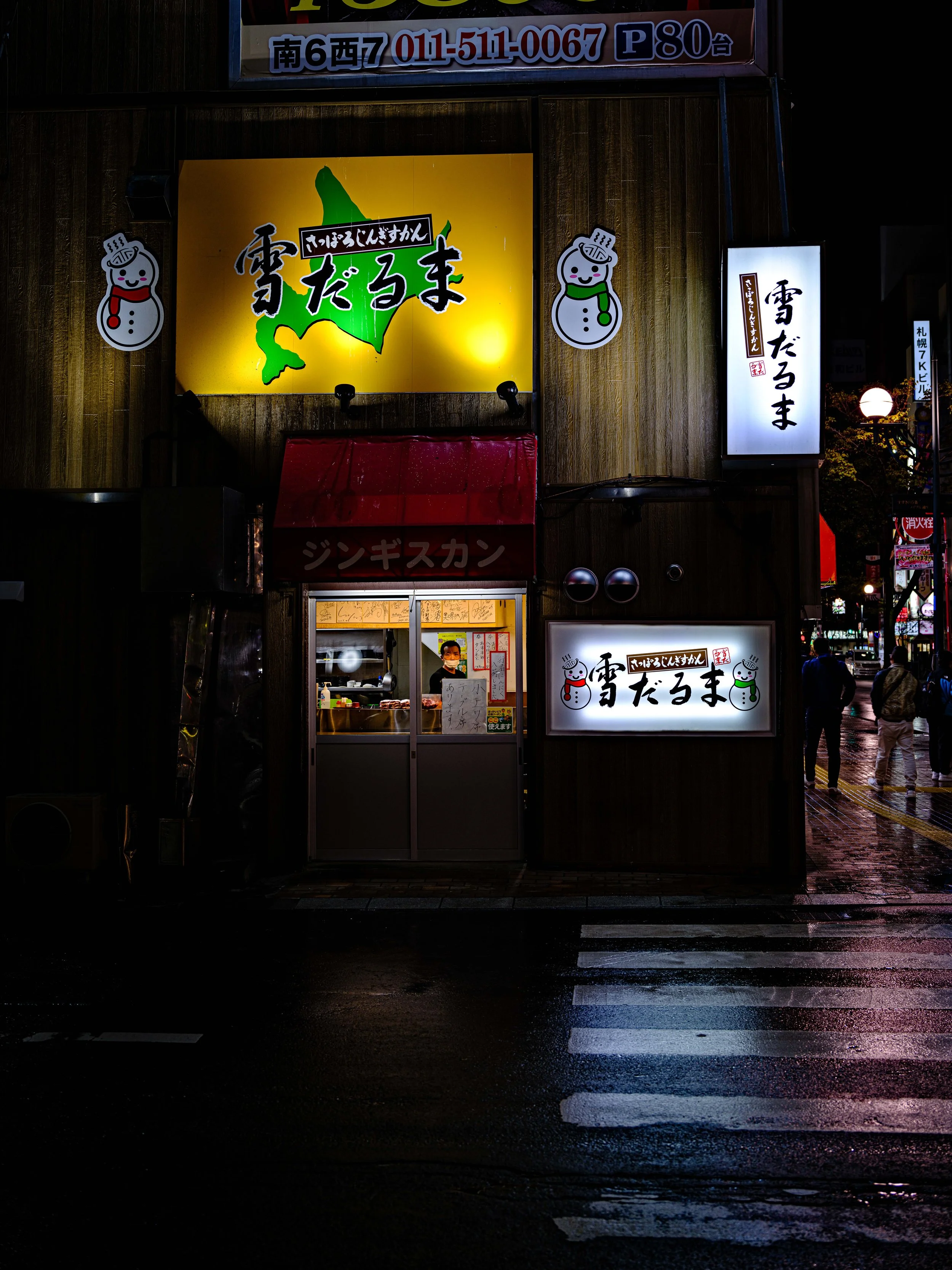 Night view of a Japanese restaurant with illuminated signs and snowman decorations, located on a street corner with wet pavement and street crosswalk markings.