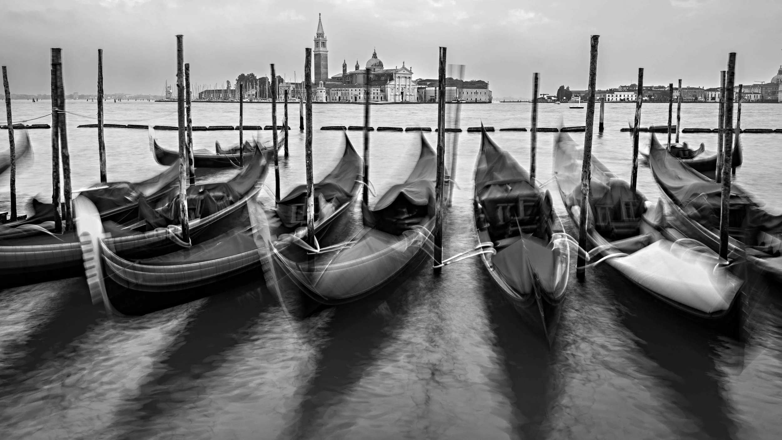 Black and white photo of several gondolas docked along a canal in Venice, Italy, with historic buildings in the background.