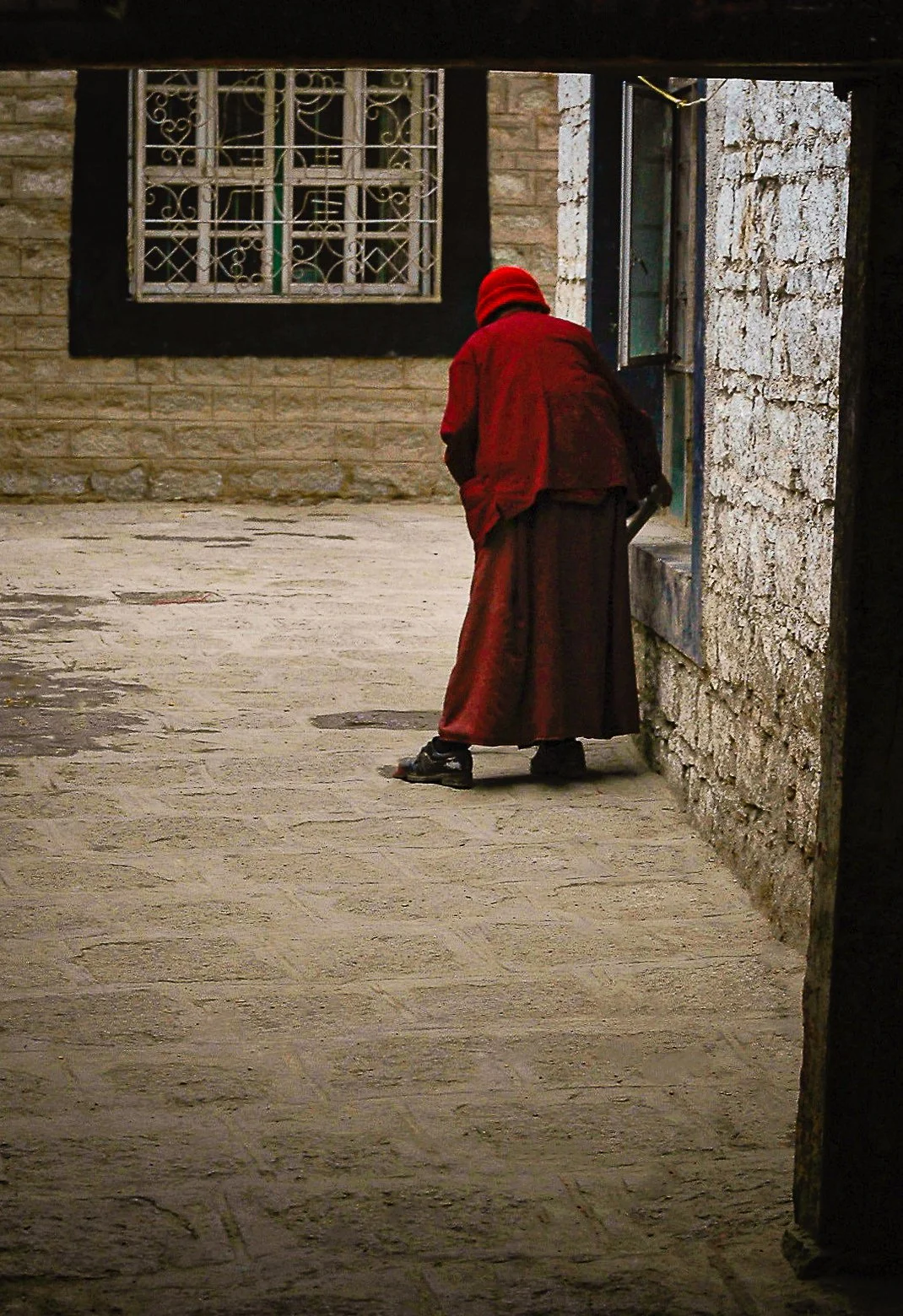 An elderly person dressed in a red coat and hat leaning against a stone wall, looking into a building through an open window.