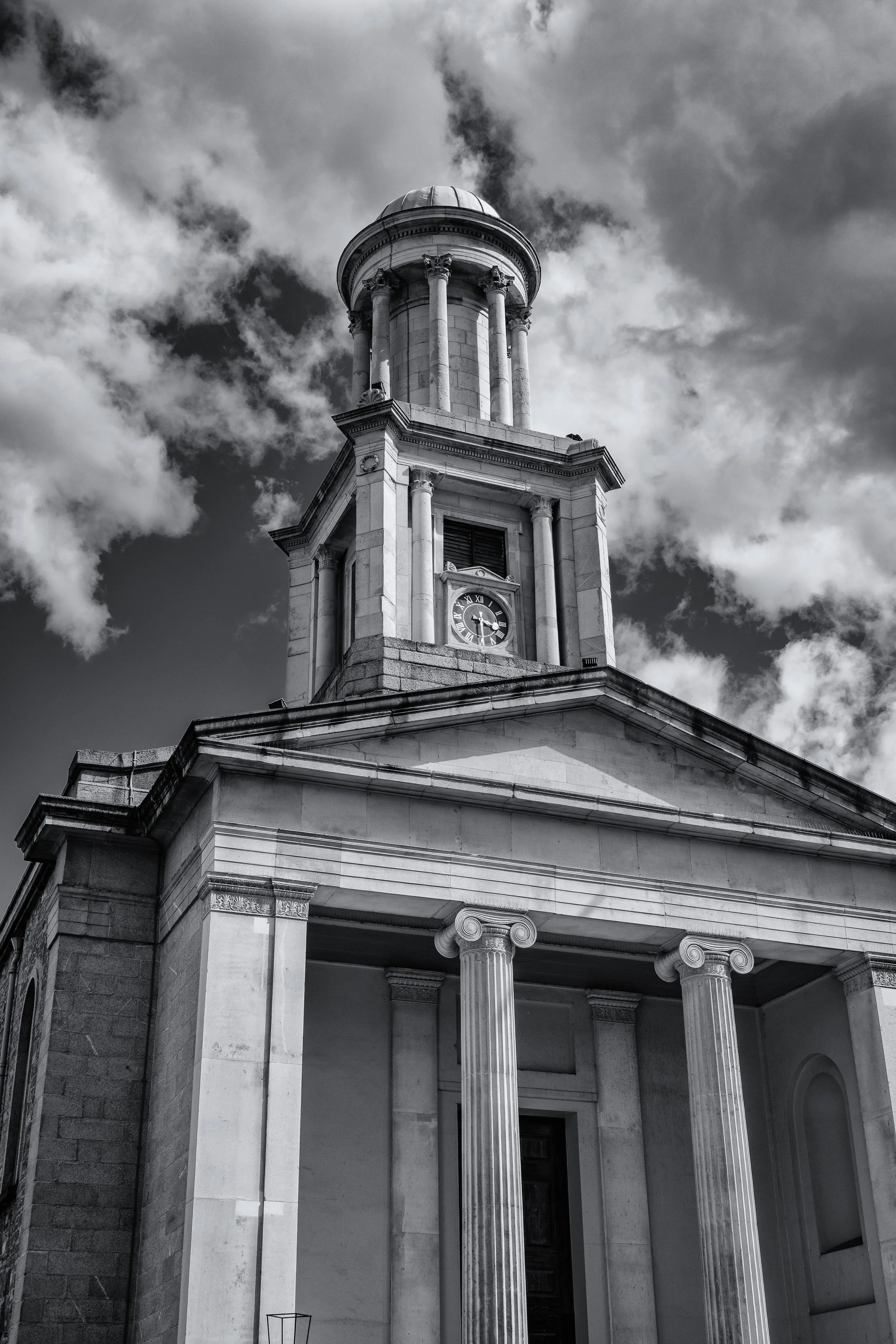 Black and white photo of a classic neoclassical building with tall columns and a clock tower, under a cloudy sky.