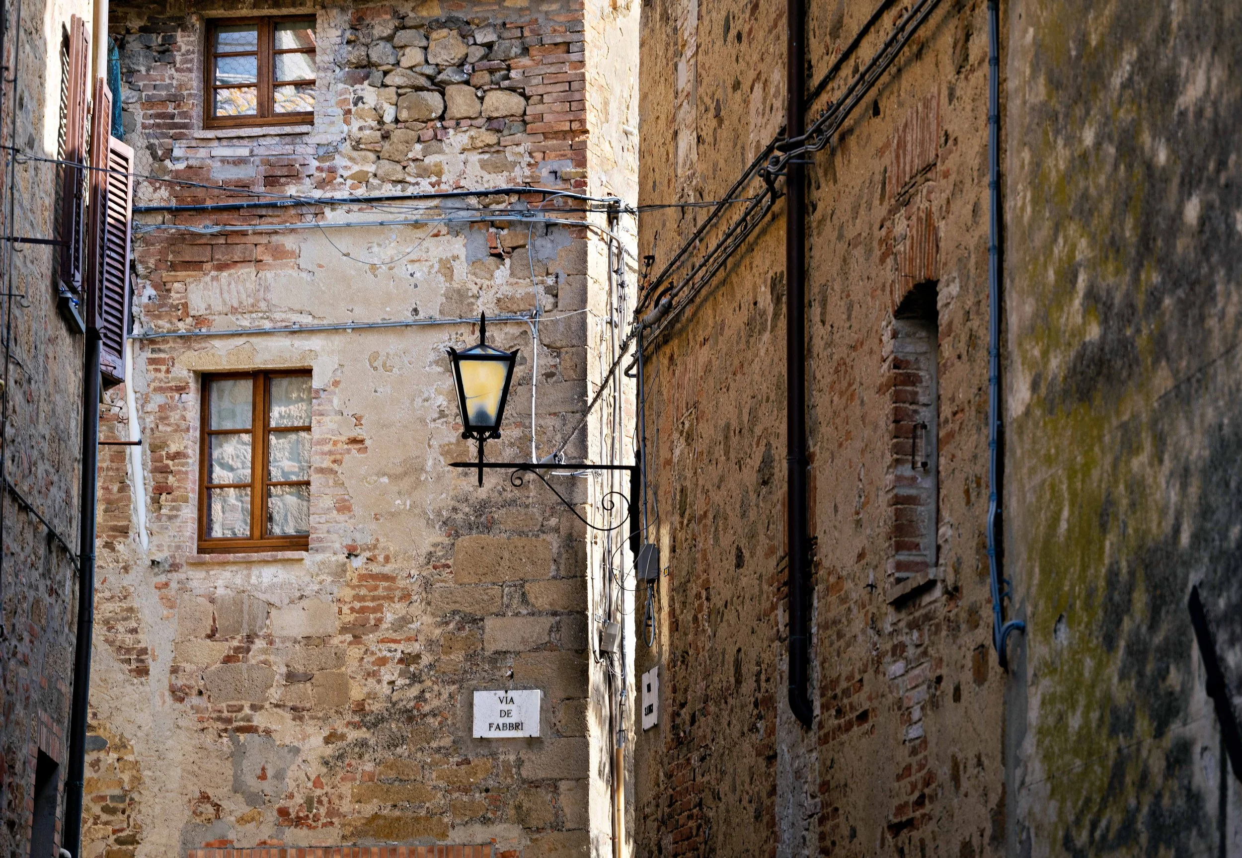 Narrow alleyway with old brick and stone walls, a streetlamp, electrical wires, and a small sign that reads "VIA DE FABBRI."