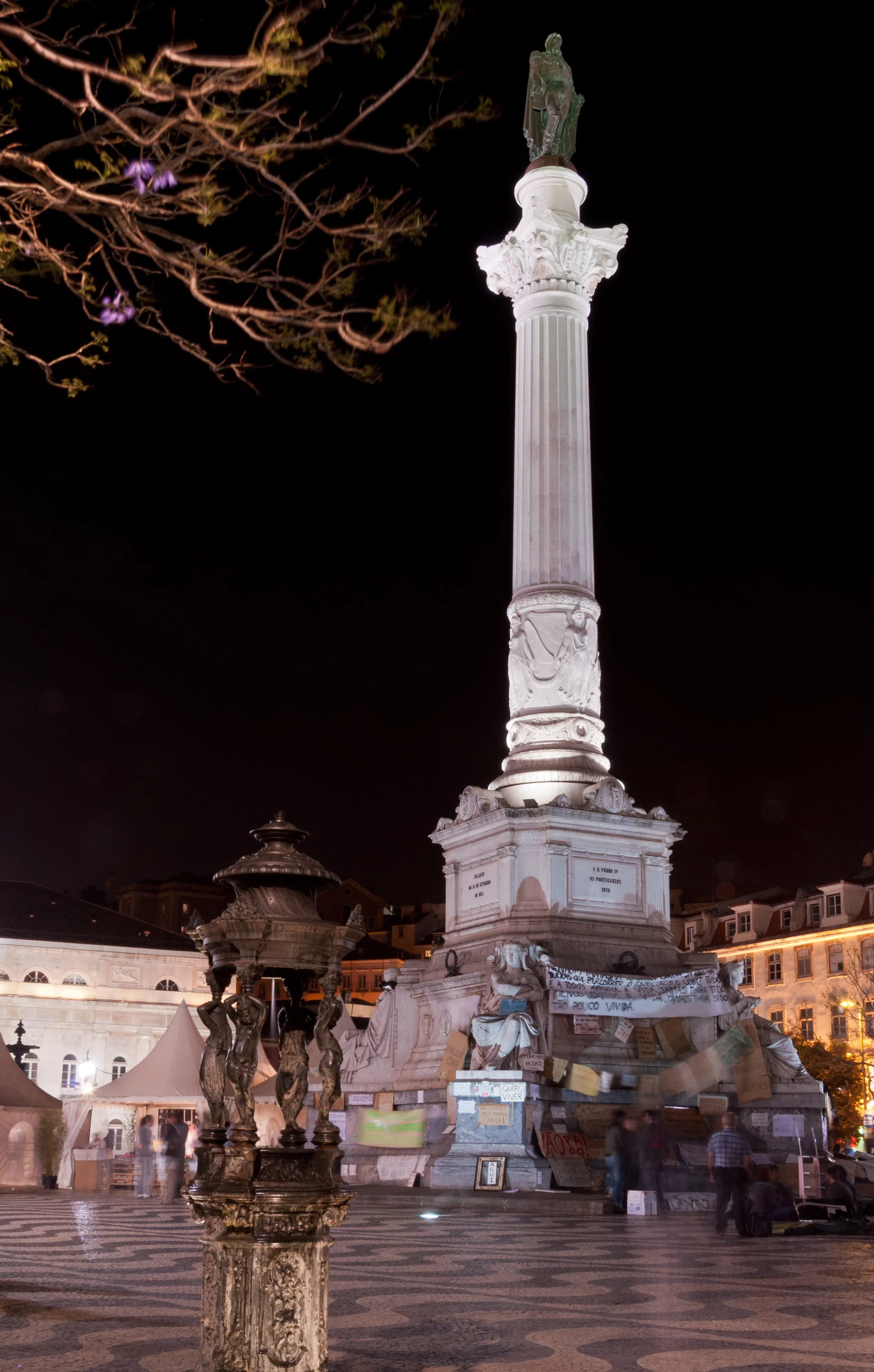 Night view of a monument with a tall column and a statue on top, situated in a decorated public square with historic buildings and a fountain, illuminated against the dark sky.