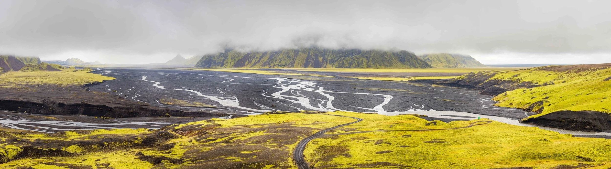 A vast landscape featuring green moss-covered hills, a winding road, and a river delta surrounded by mountains with low clouds overhead.