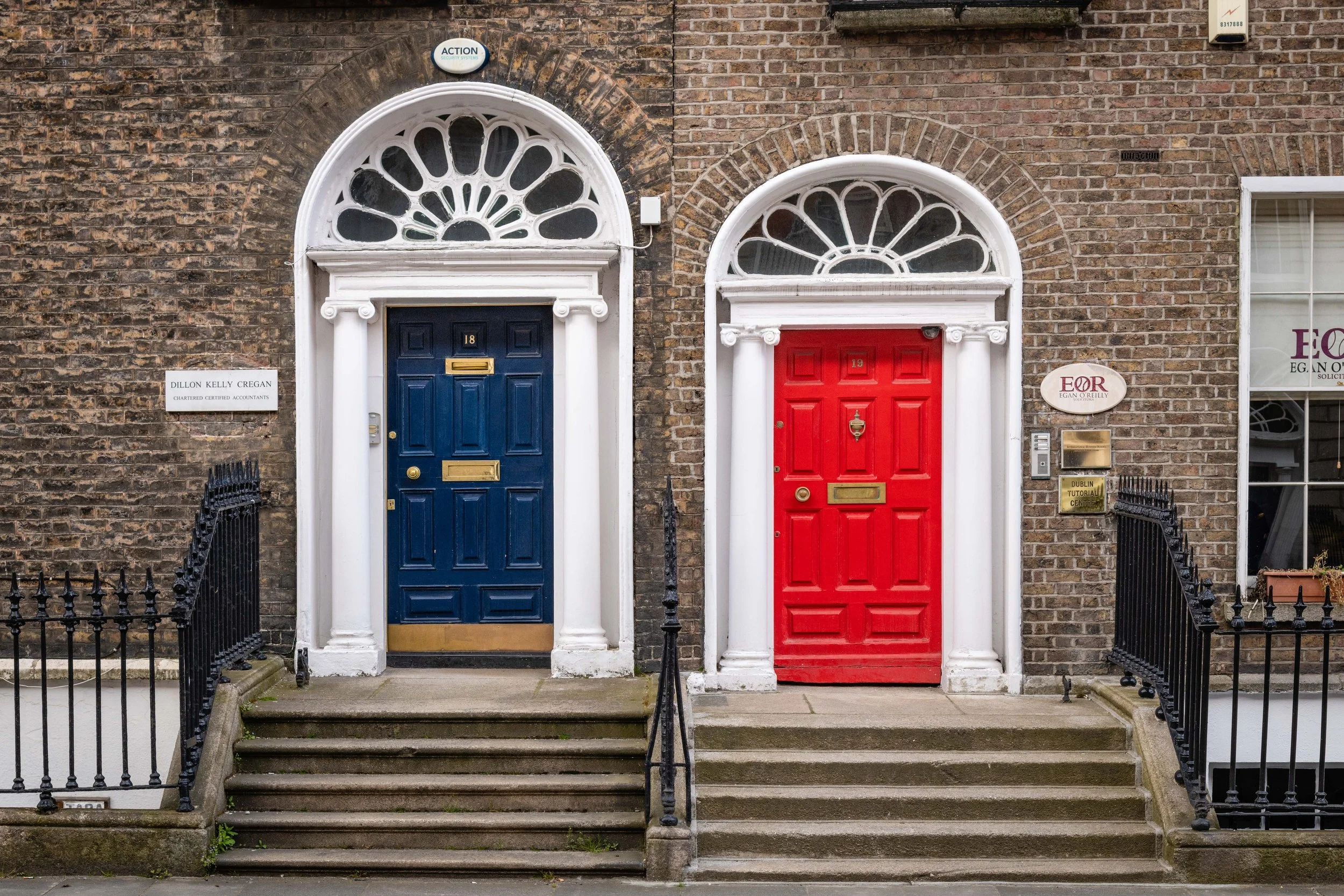 Two colorful front doors, one blue and one red, with white decorative archways and steps, on a brick building with various signs.
