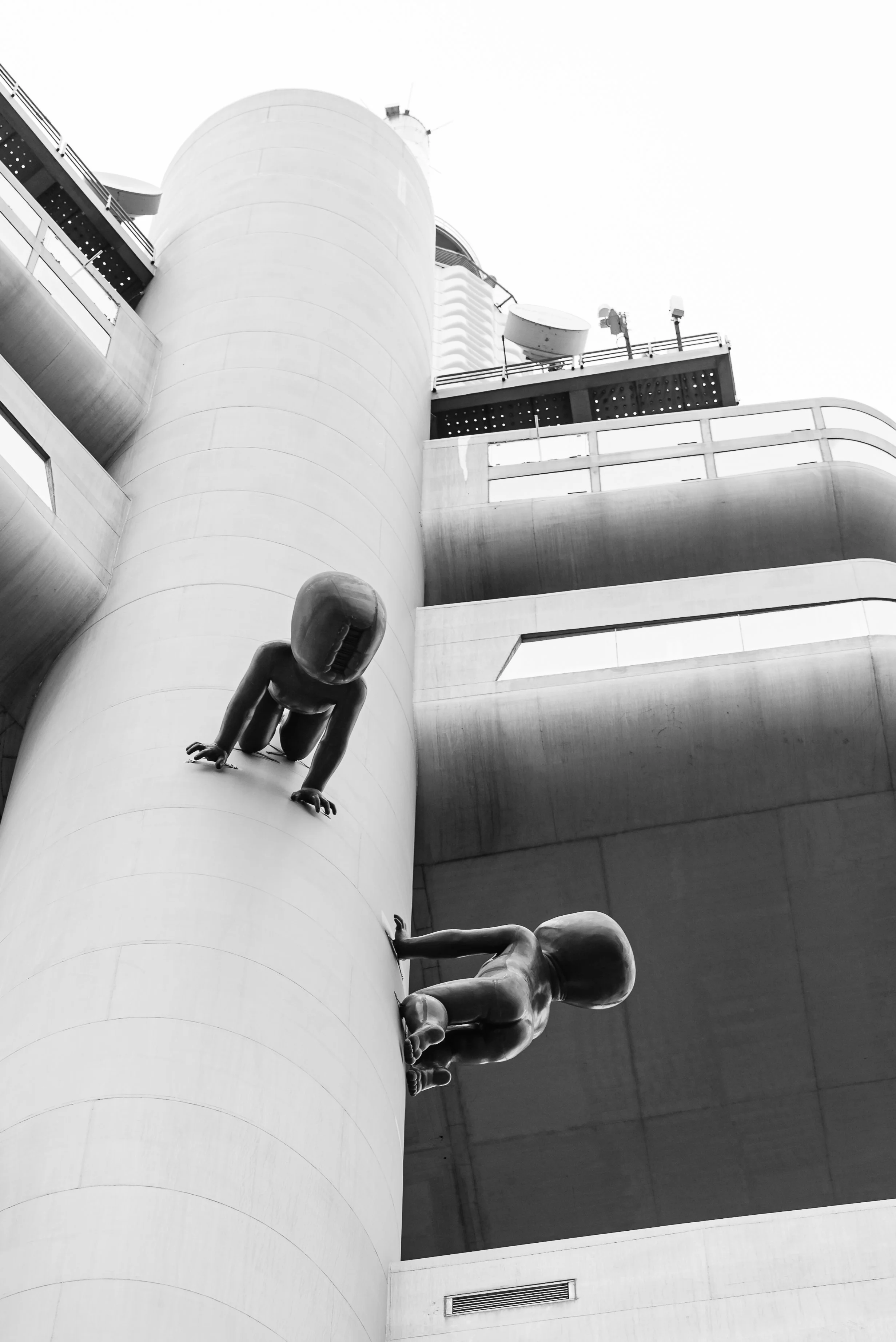 Black and white photo of an architectural structure with two modern child sculptures climbing a cylindrical wall, with one sculpture appearing to be a crawling baby and the other a toddler, set against a geometric building background.