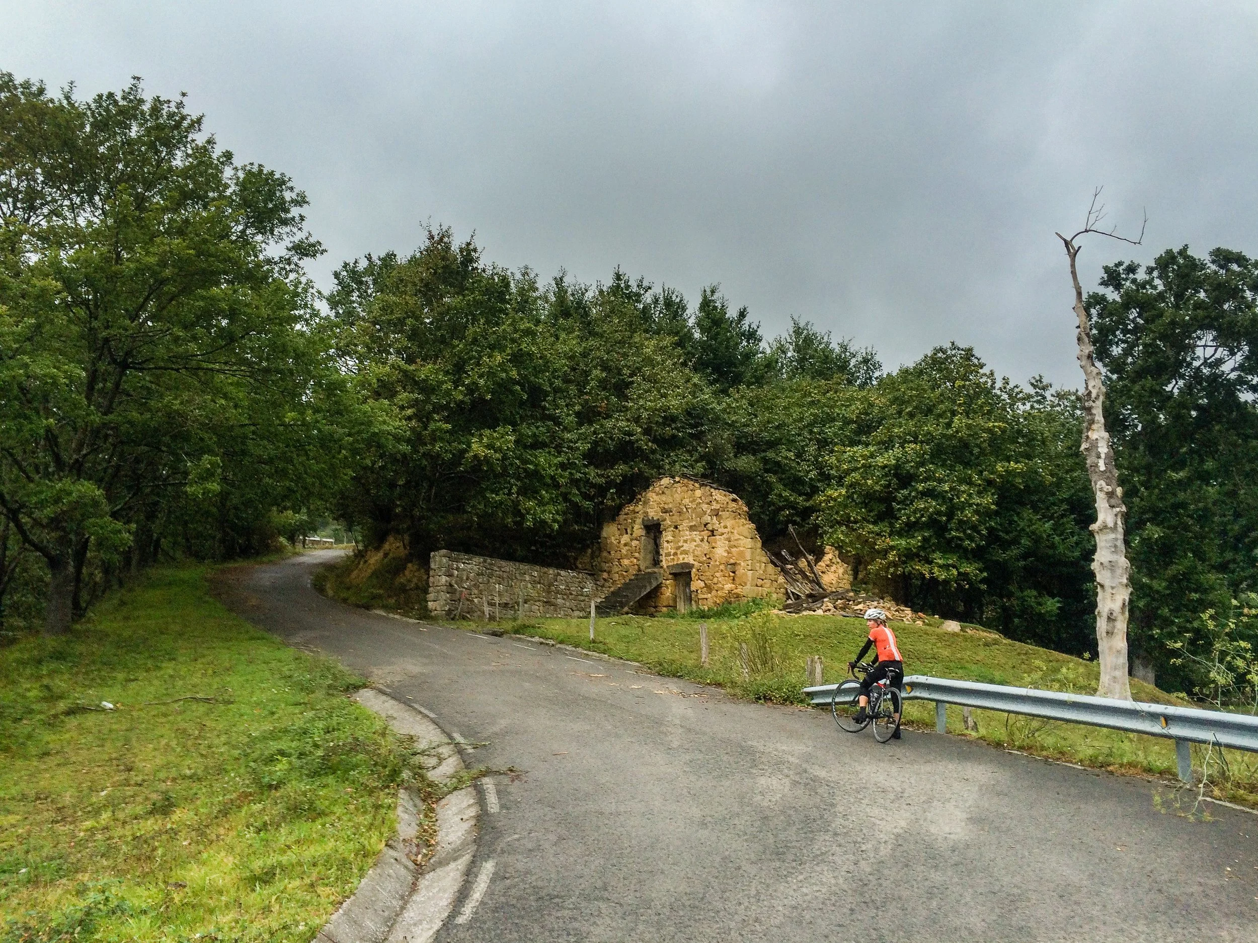 A cyclist rides on a narrow, winding road past an abandoned stone building surrounded by trees, with a cloudy sky overhead.