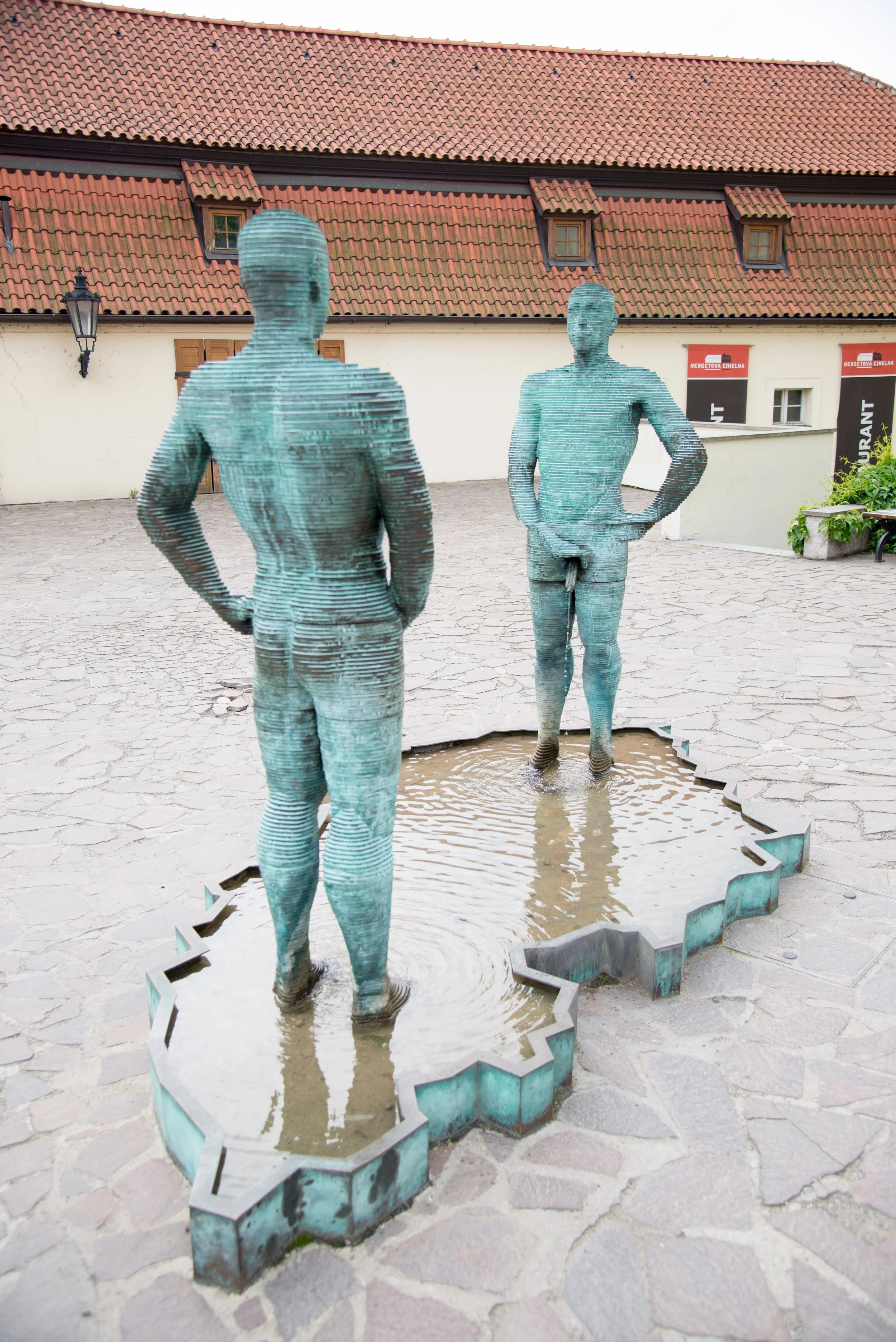 Two bronze statues of men standing in a shallow water feature outdoors, facing each other, with a building with a red-tiled roof in the background.