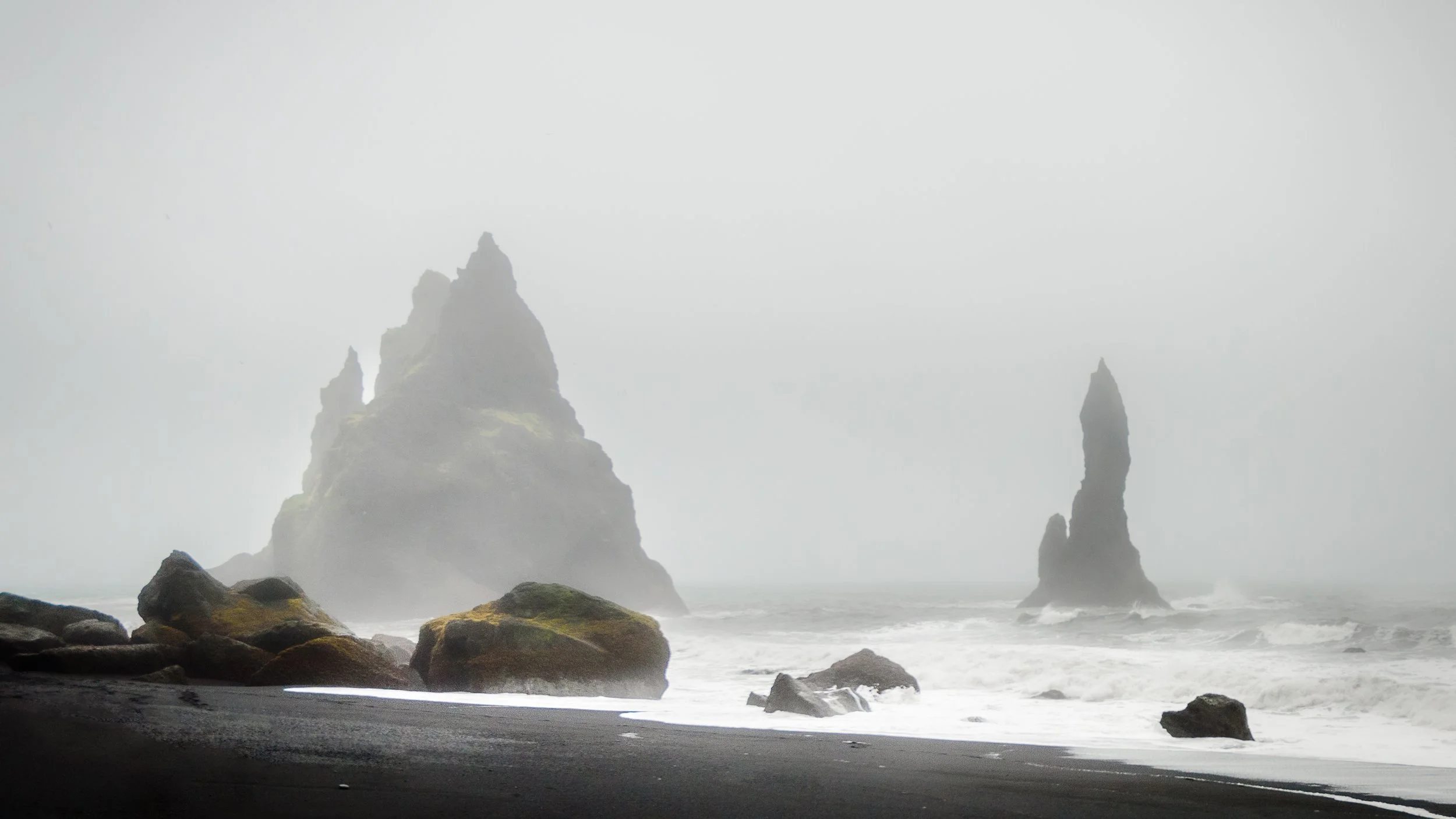 Misty black sand beach with large rocks in the foreground and tall sea stacks emerging from foggy ocean in the background.