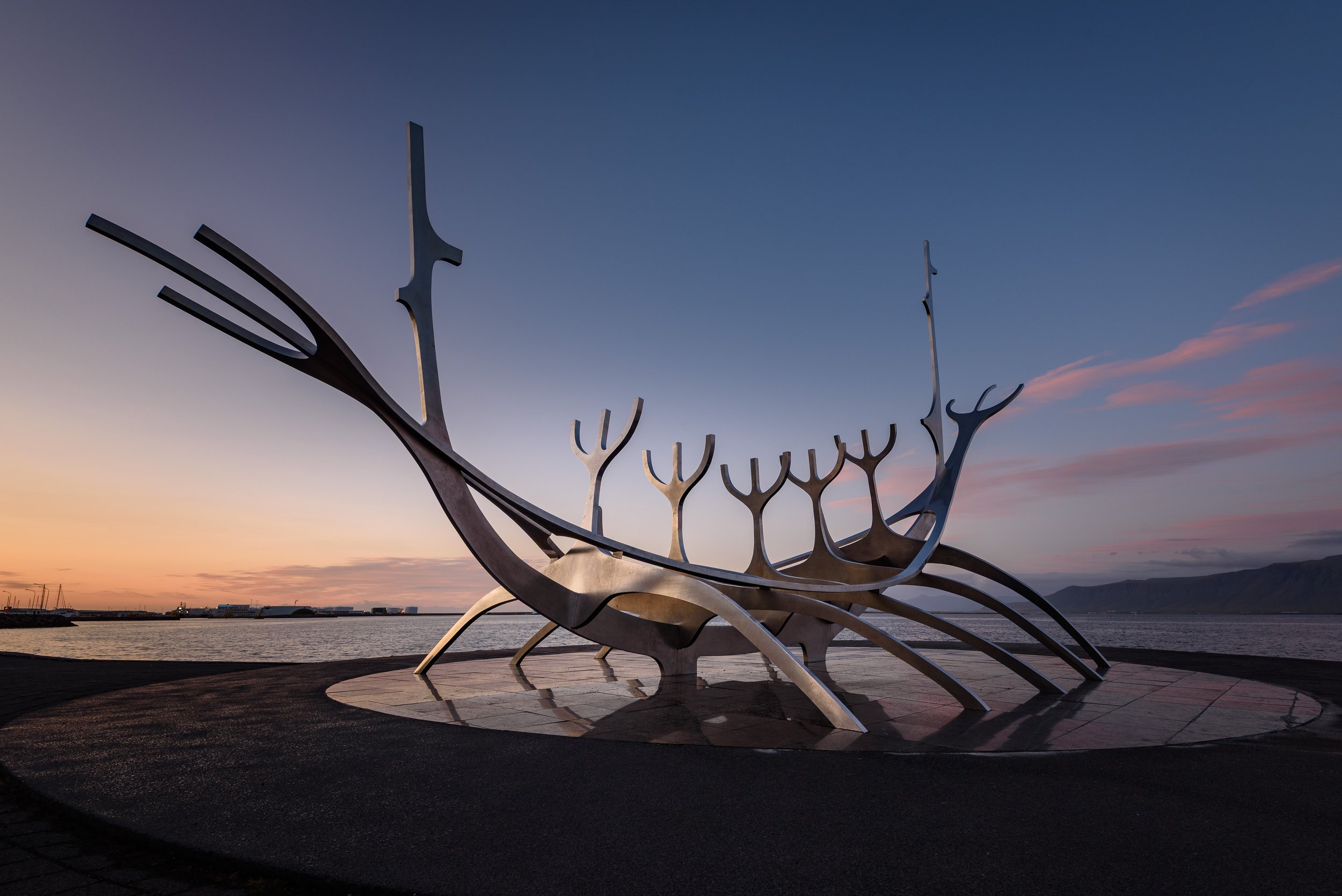 A modern sculpture of a Viking ship with three tall, curved horns on each side, situated near a body of water at sunset, with mountains visible in the distance.