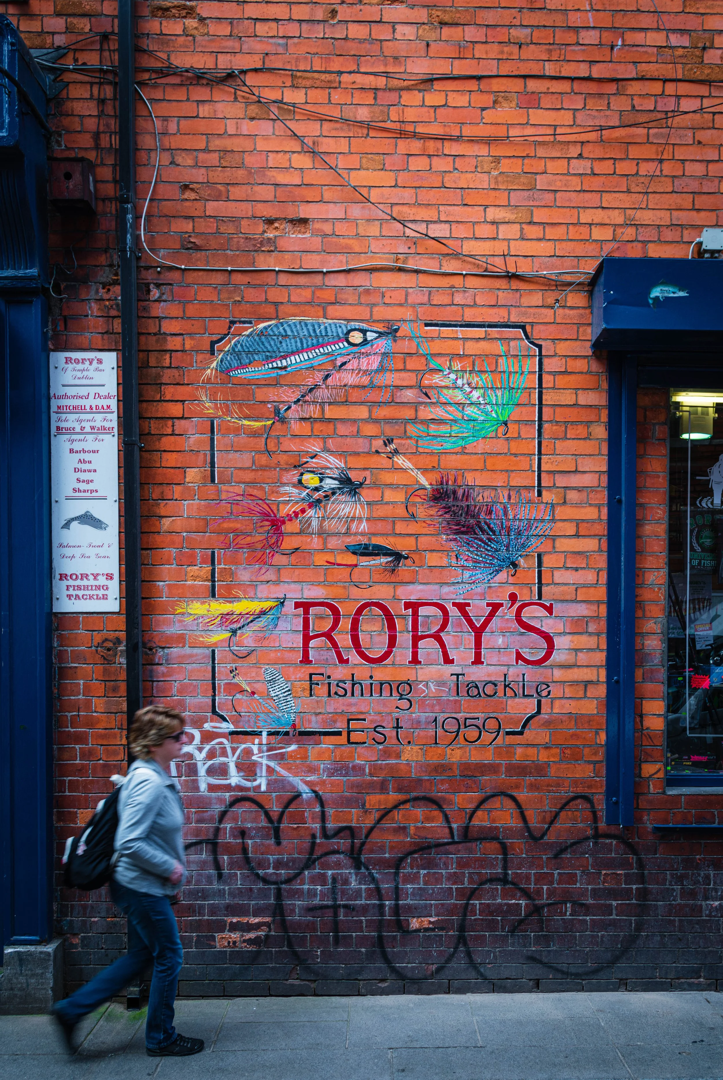 A brick wall with a colorful mural advertising Rory's Fishing Tackle, established in 1959, featuring painted images of fishing lures and fish. A person with a backpack walks past on the sidewalk.