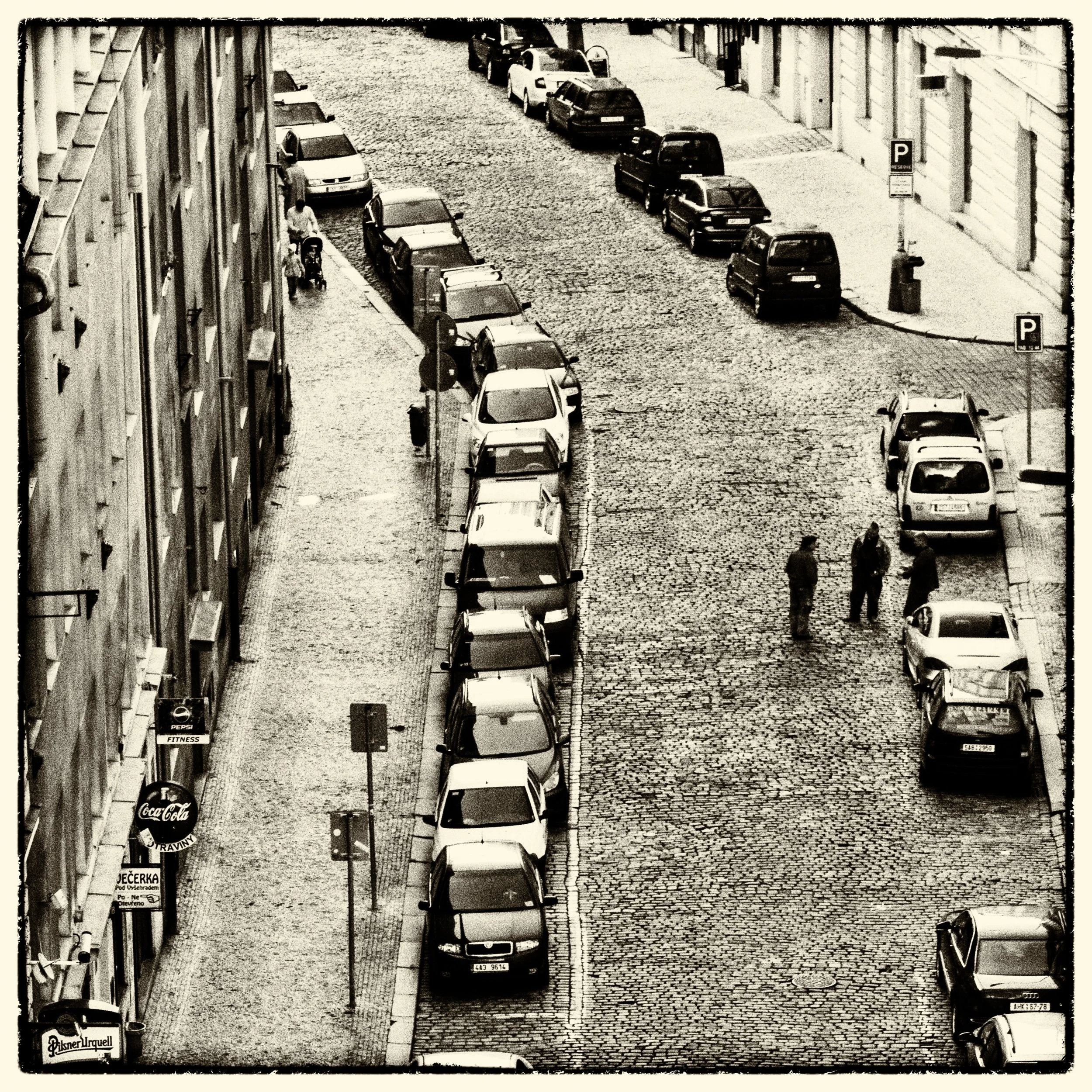 Black and white photo of a cobblestone street with parked cars lining the sides, a few people standing and talking, buildings on both sides, and storefront signs visible.