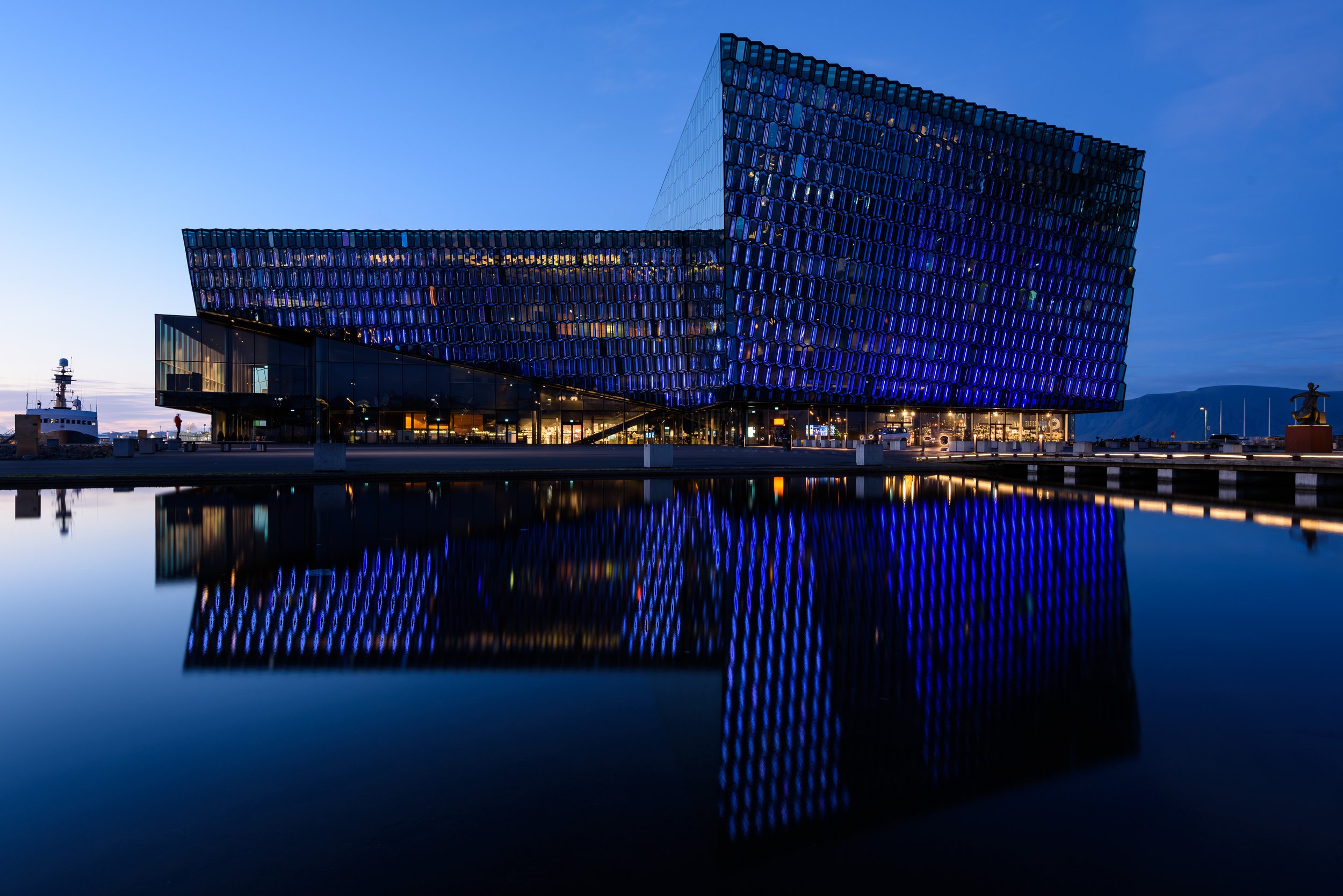 A modern, blue-lit building with geometric design, reflected in a body of water at dusk.