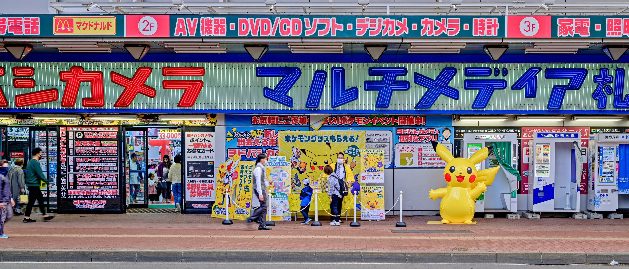 Exterior of a Japanese electronics and camera store decorated with Pikachu themed signs and a large Pikachu inflatable outside, with several people entering and exiting.