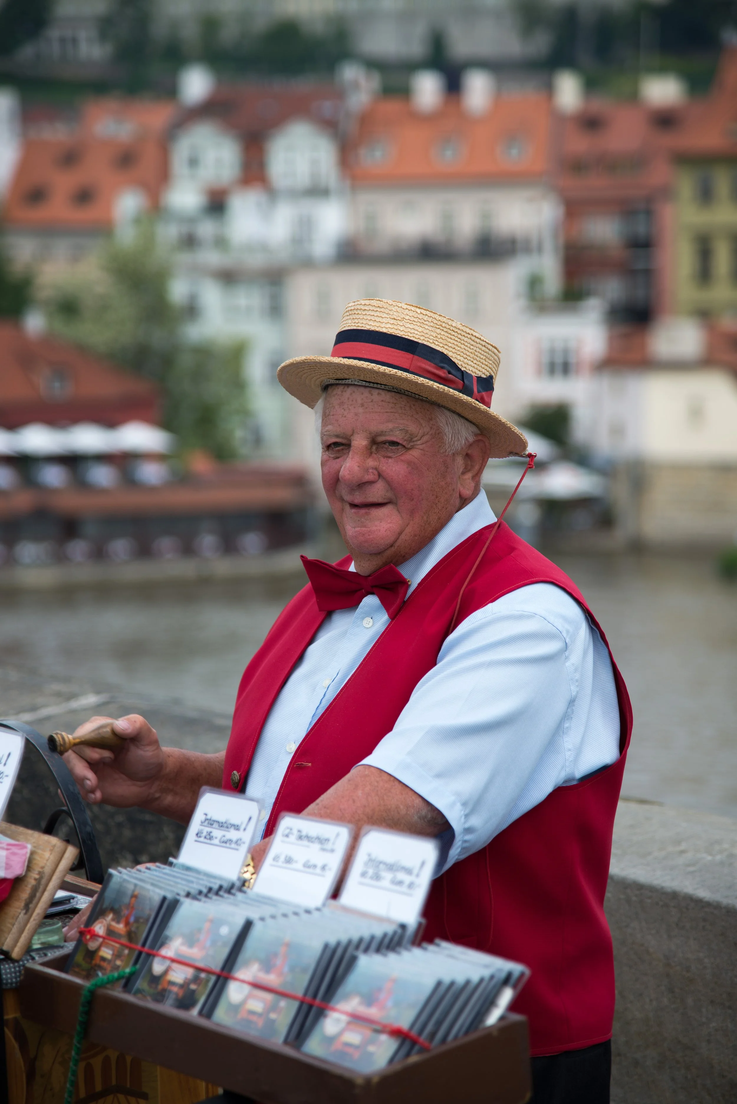 An older man dressed as a traditional souvenir vendor, wearing a red vest, a red bow tie, and a straw boater hat, standing by a market stall with postcards, with a river and colorful historic buildings in the background.