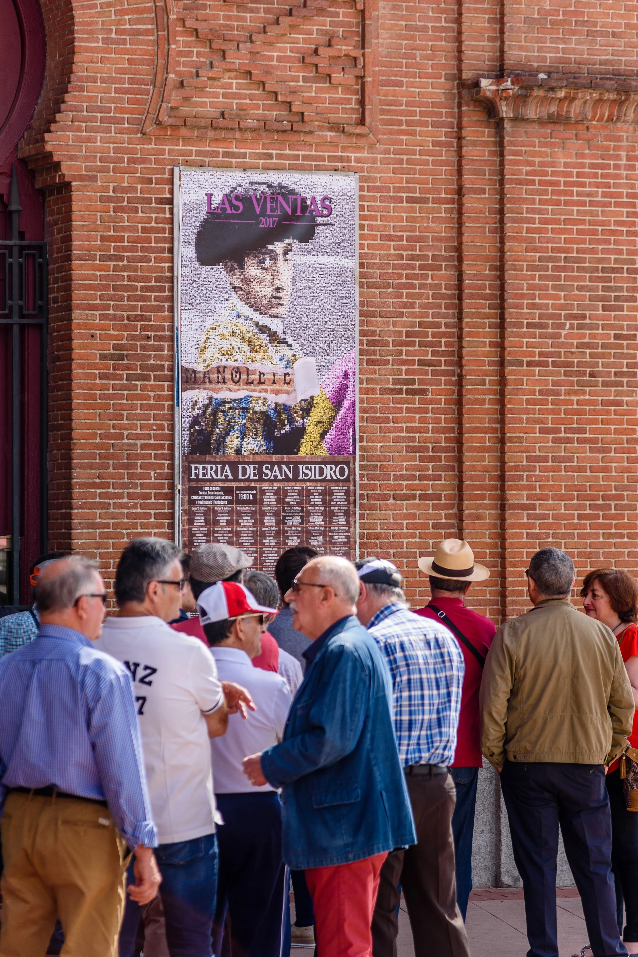 Group of people gathered in front of a brick wall with a poster of a woman wearing traditional attire. The poster advertises the Las Ventas 2017 event at Feria de San Isidro.