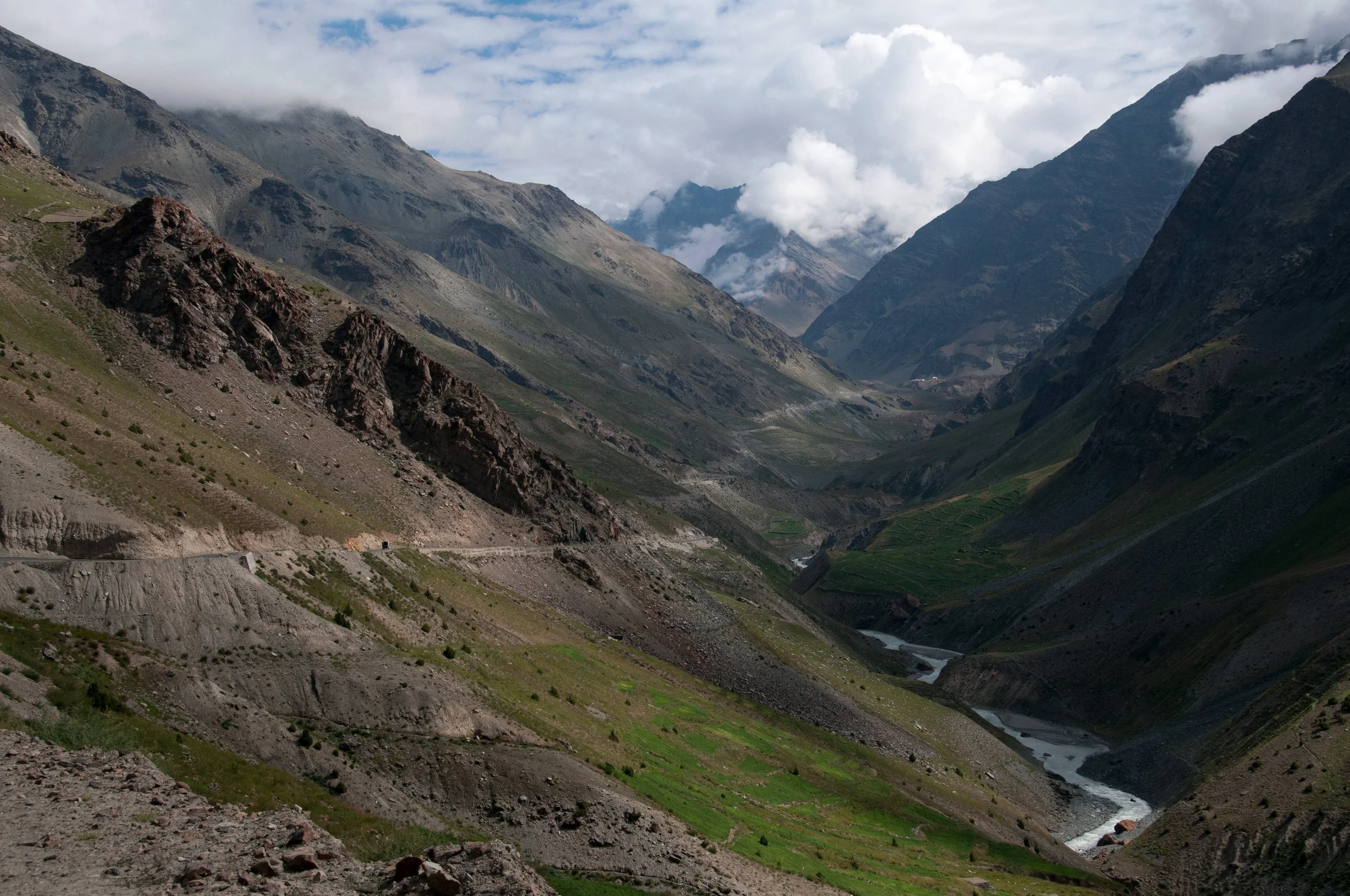 Mountain landscape with steep slopes, a winding river, and cloudy sky.