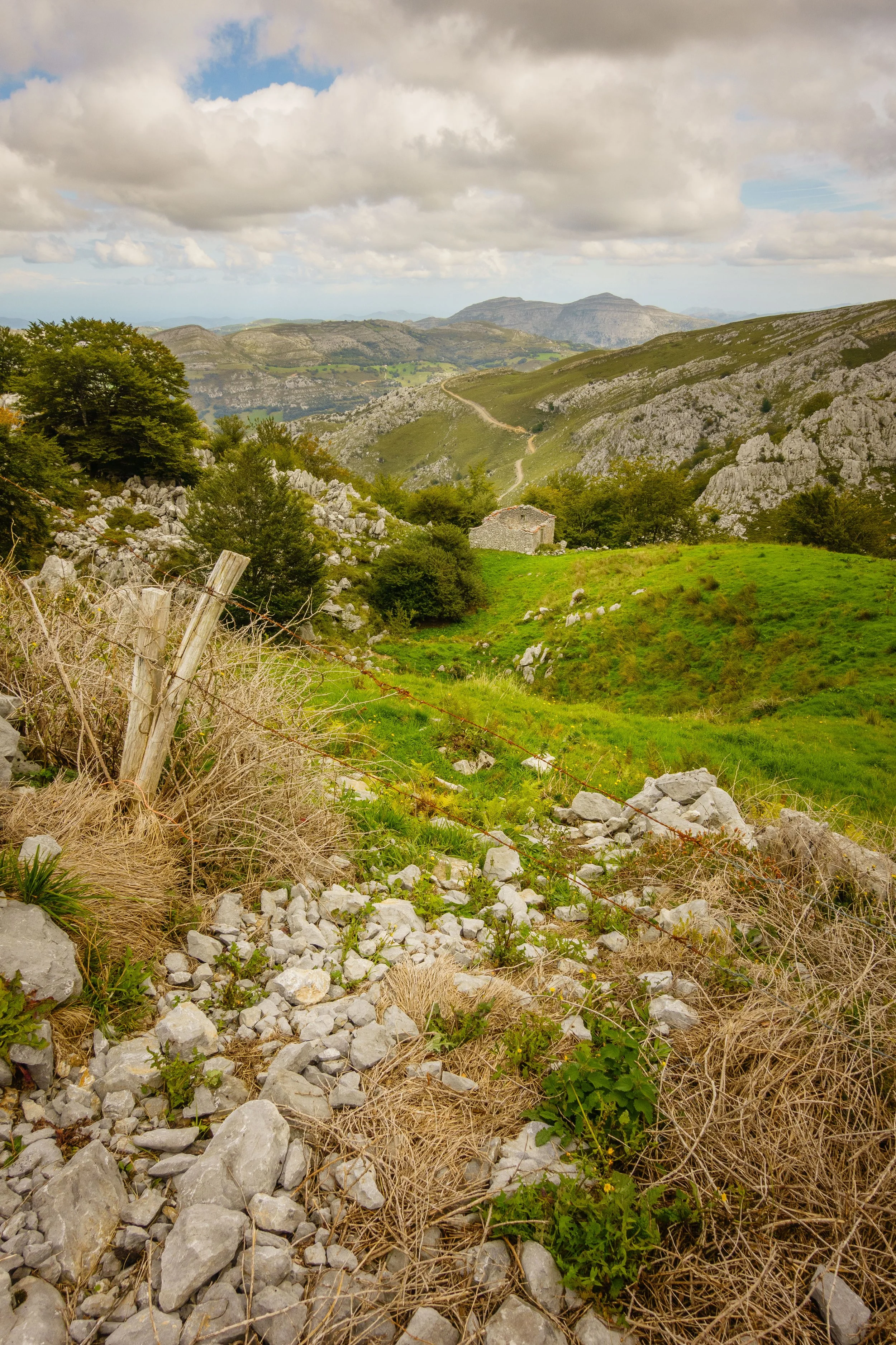 A landscape of rolling green hills, rocky terrain, and a cloudy sky, with a stone building in the middle distance and a dirt path winding through the hills.