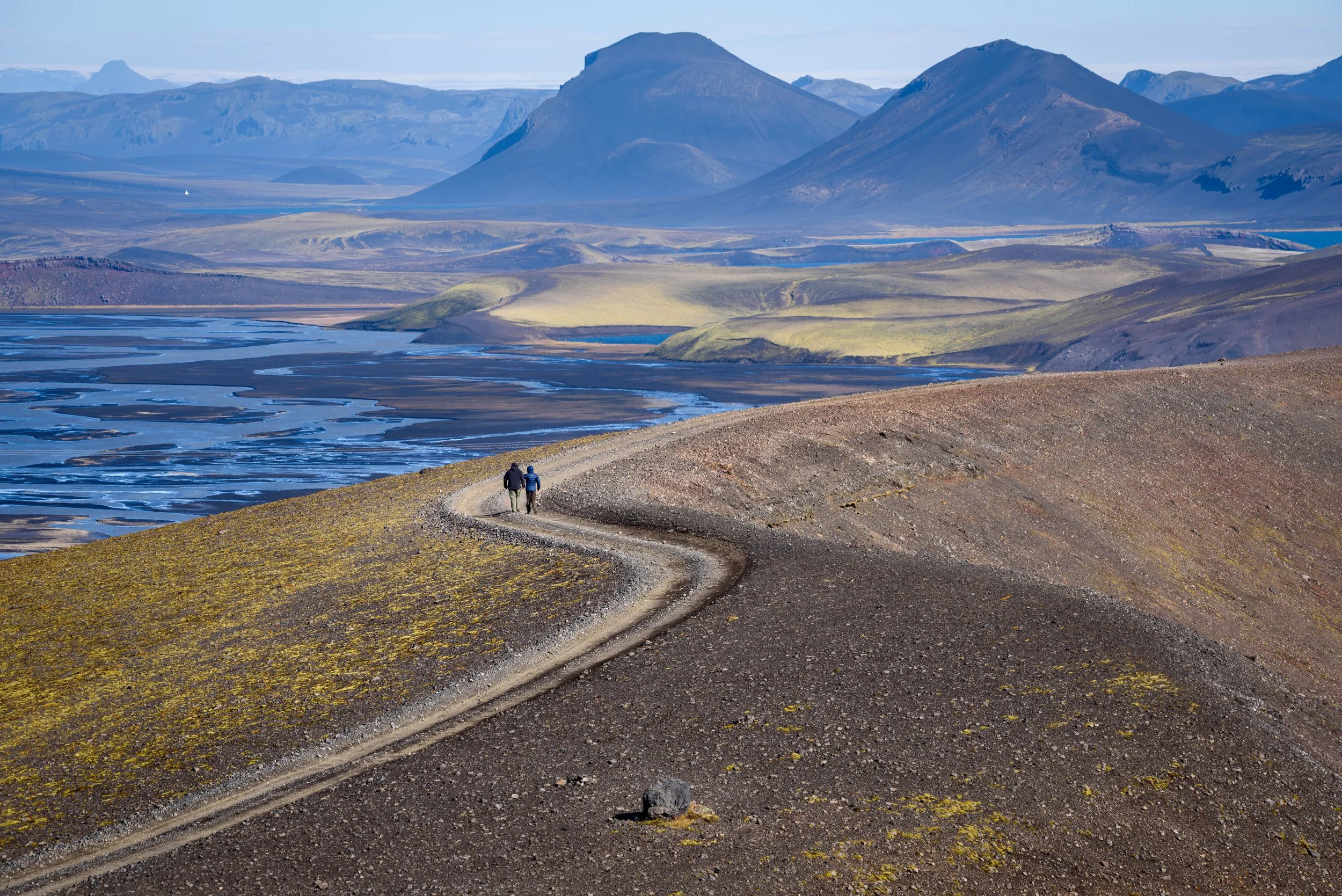 Two hikers walking along a winding dirt trail in a volcanic landscape with distant mountains and a lake.