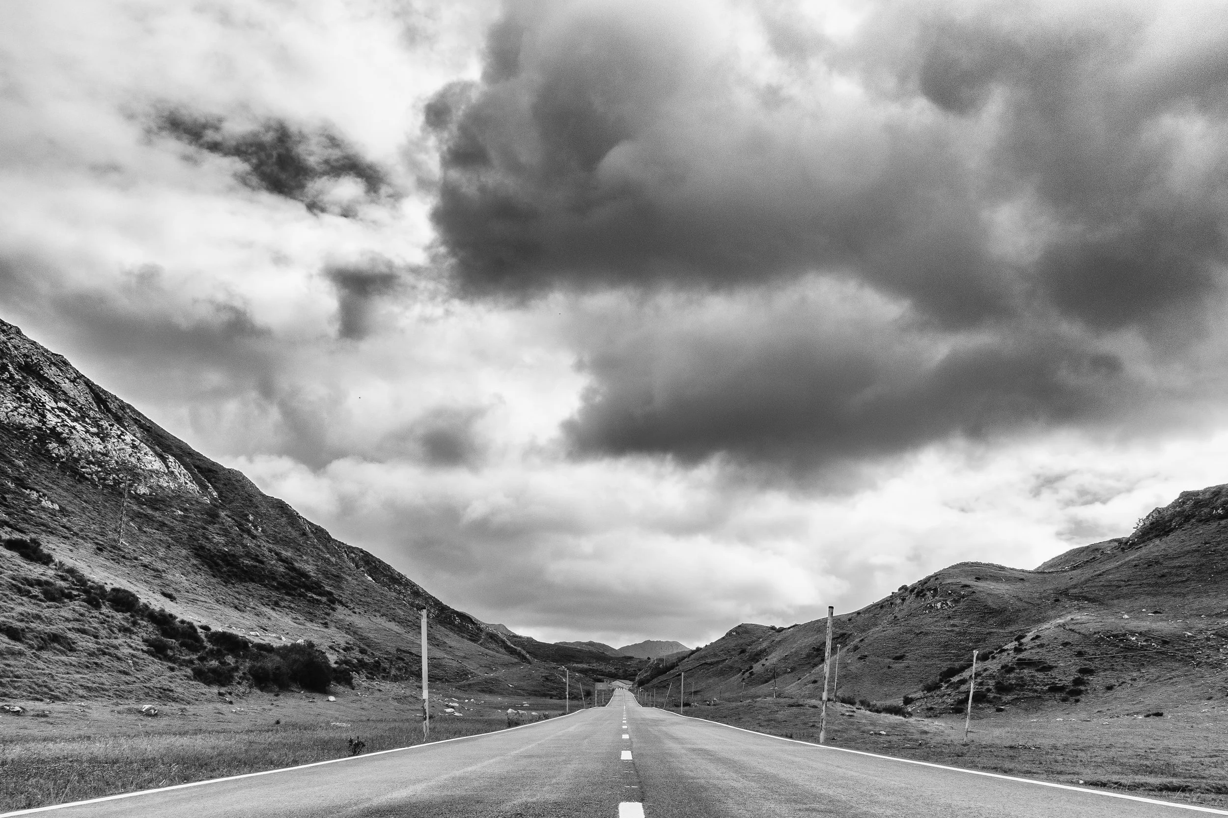 Black and white photo of an empty long straight road extending into the distance between mountains with cloudy sky overhead.