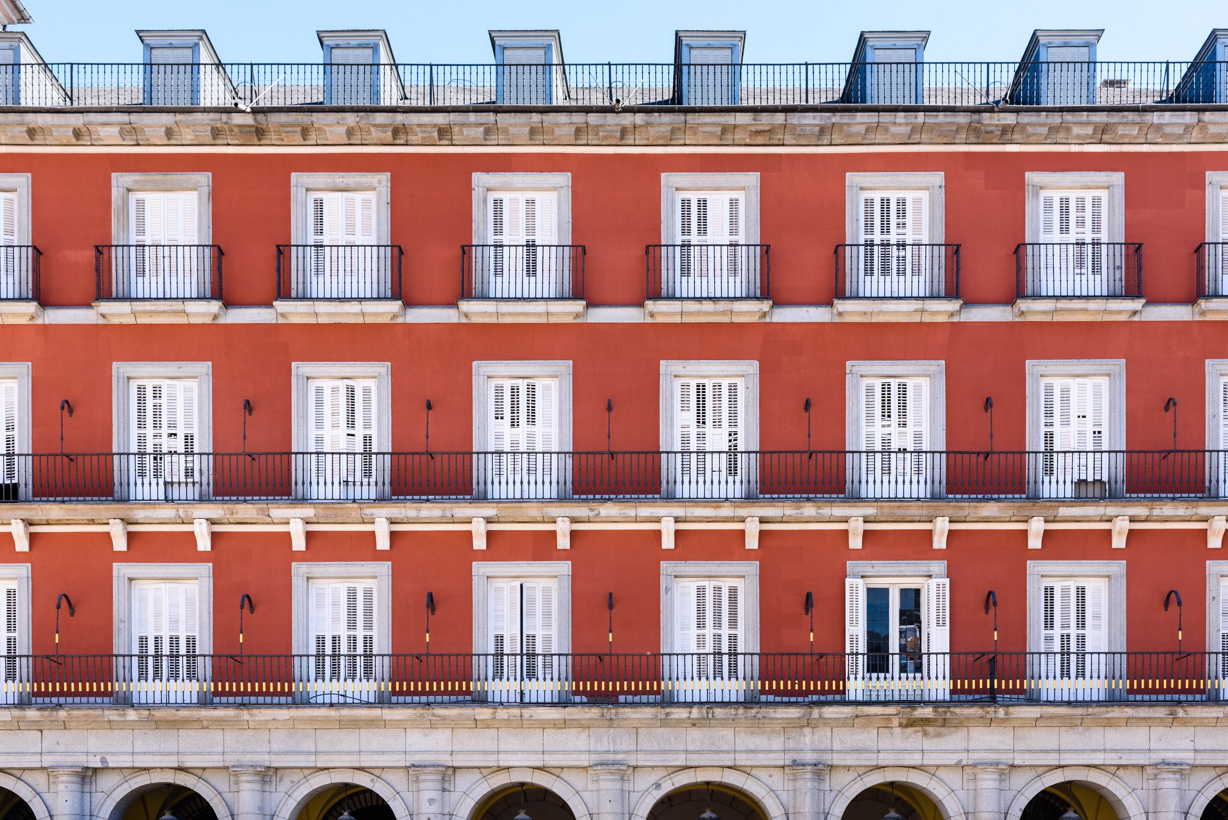 Multi-story yellow building with a red facade, white window shutters, black wrought-iron balconies, and a rooftop terrace with glass skylights.