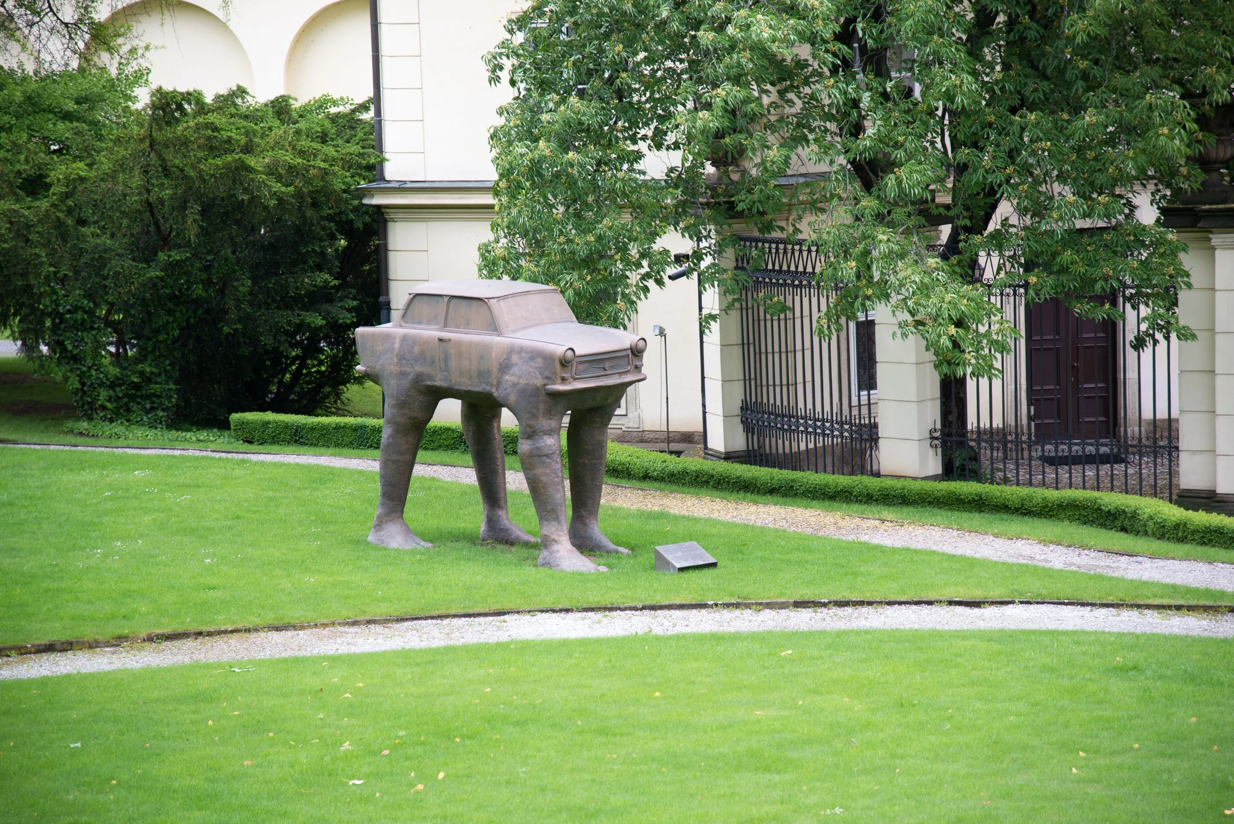 A modern sculpture of a car with four legs resembling an animal, situated on a well-maintained lawn with a pathway nearby, in front of a house with a white facade, black iron gate, and lush trees.