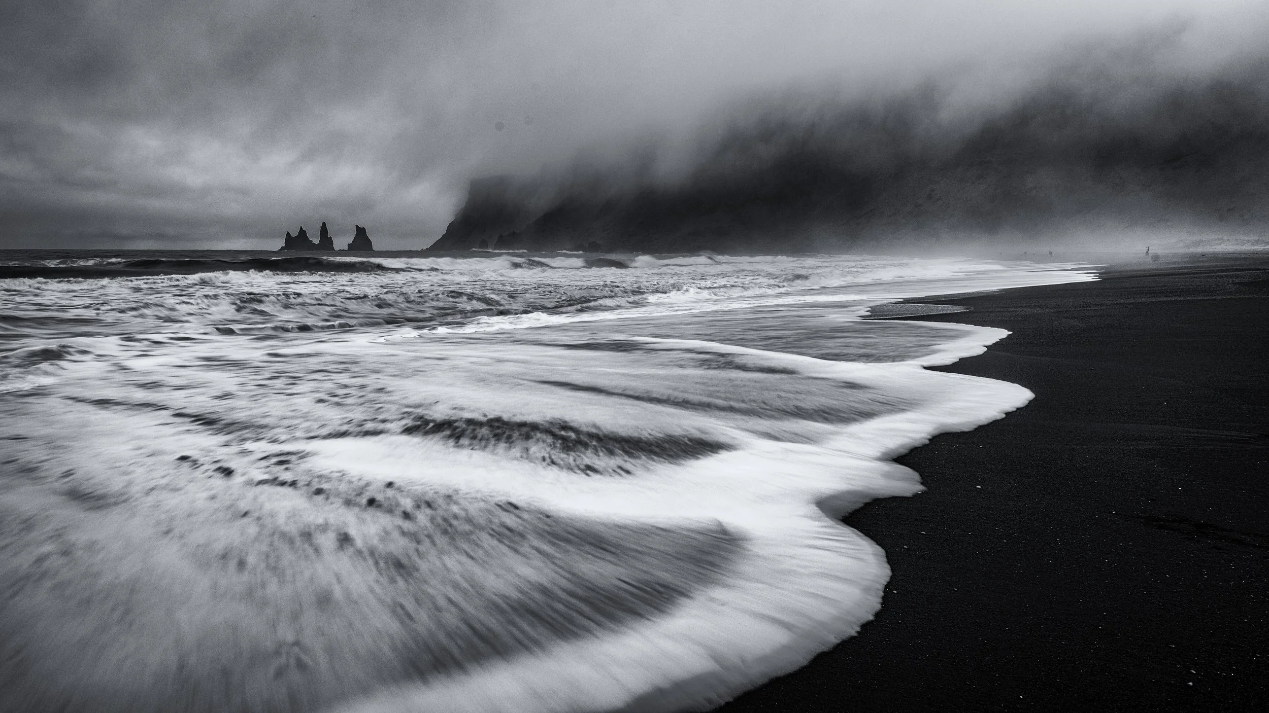 Black and white photo of a rocky beach with waves crashing on the shore and dark clouds overhead.