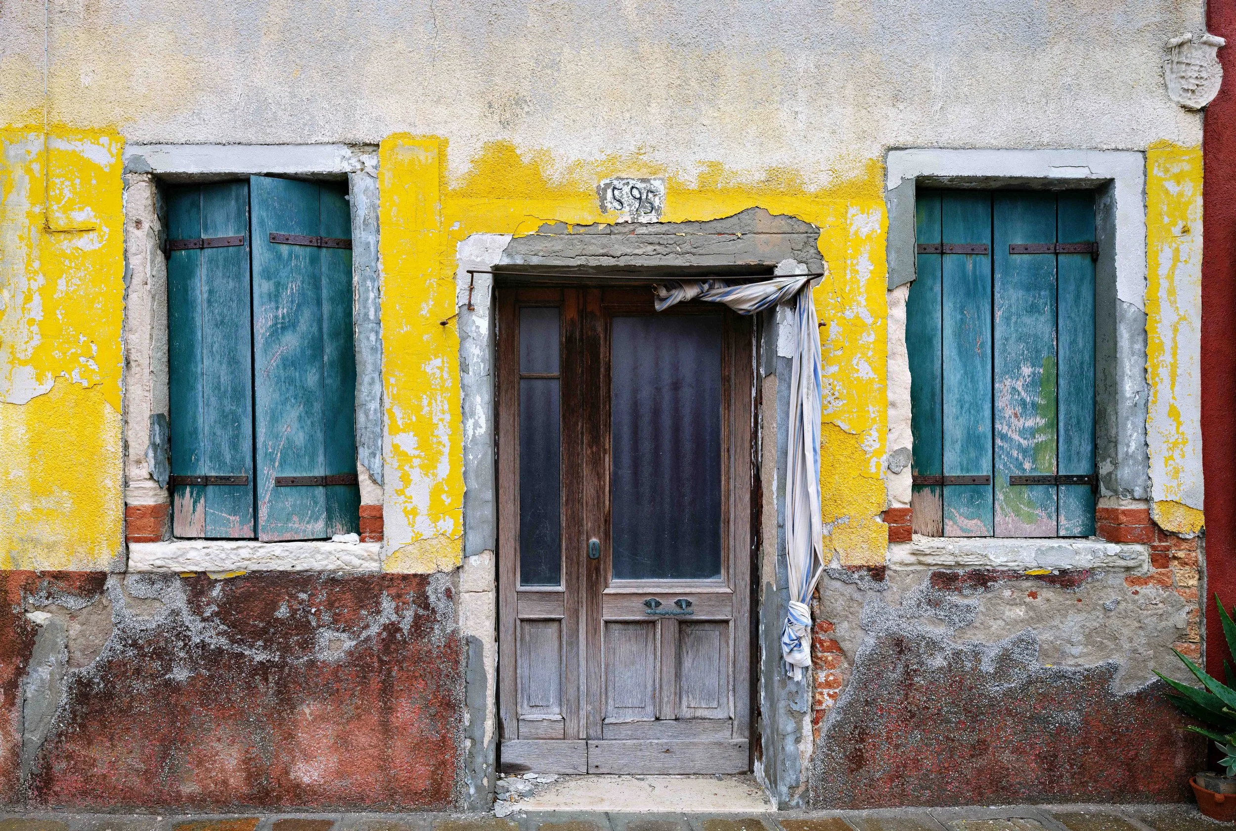 Old building facade with yellow, red, and beige peeling paint, two windows with blue shutters, and a weathered wooden door, with a striped cloth hanging by the door frame.