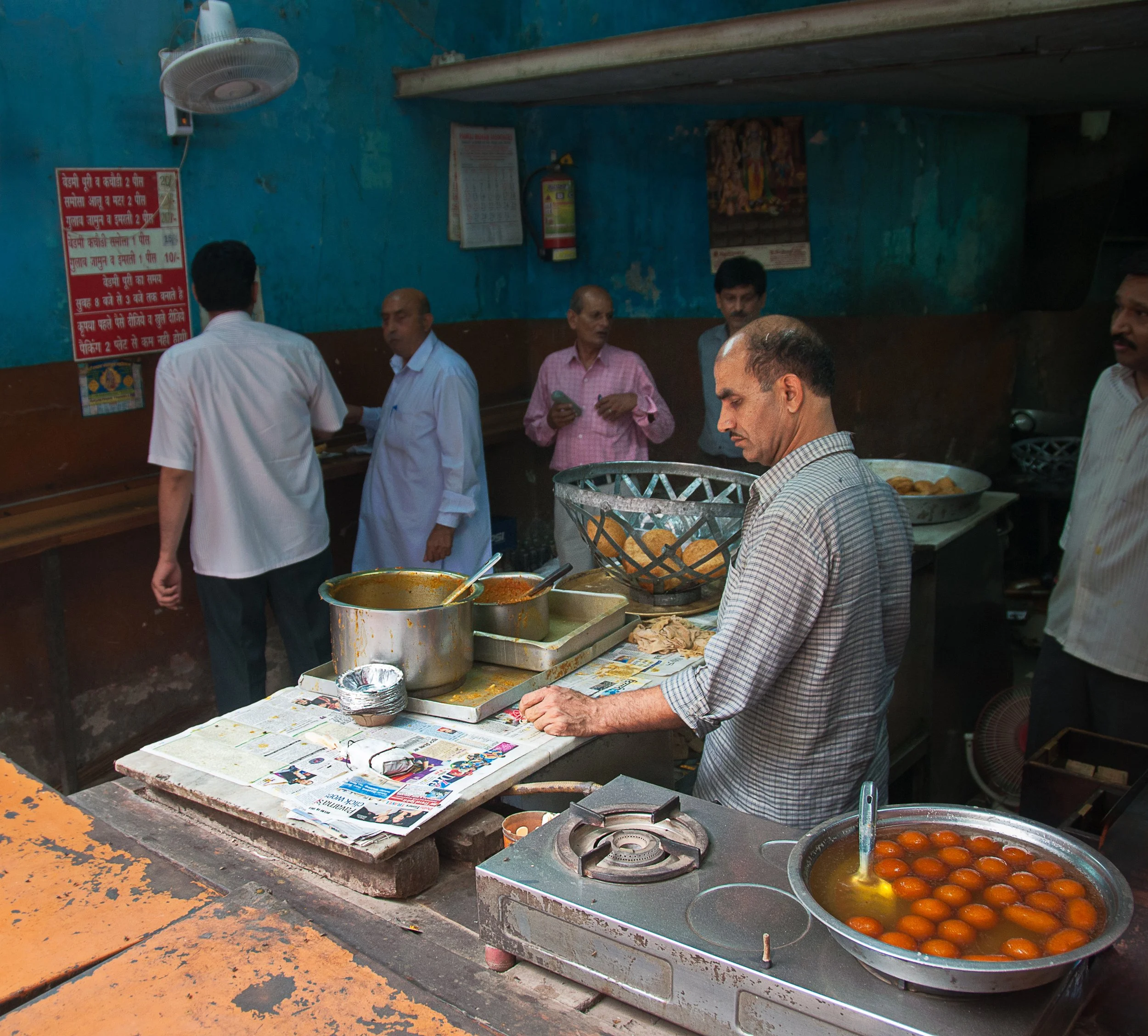 Man preparing food at a street food stall with pots of curries, a bowl of gulab jamun, and surrounded by men in a busy eatery with a blue wall.