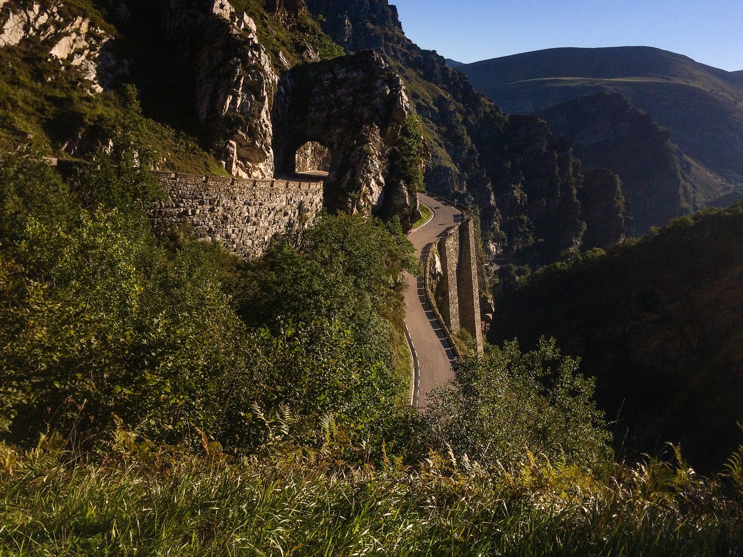 A winding mountain road with a stone barrier, surrounded by tall green trees and rocky cliffs, with hills in the distance under a clear blue sky.