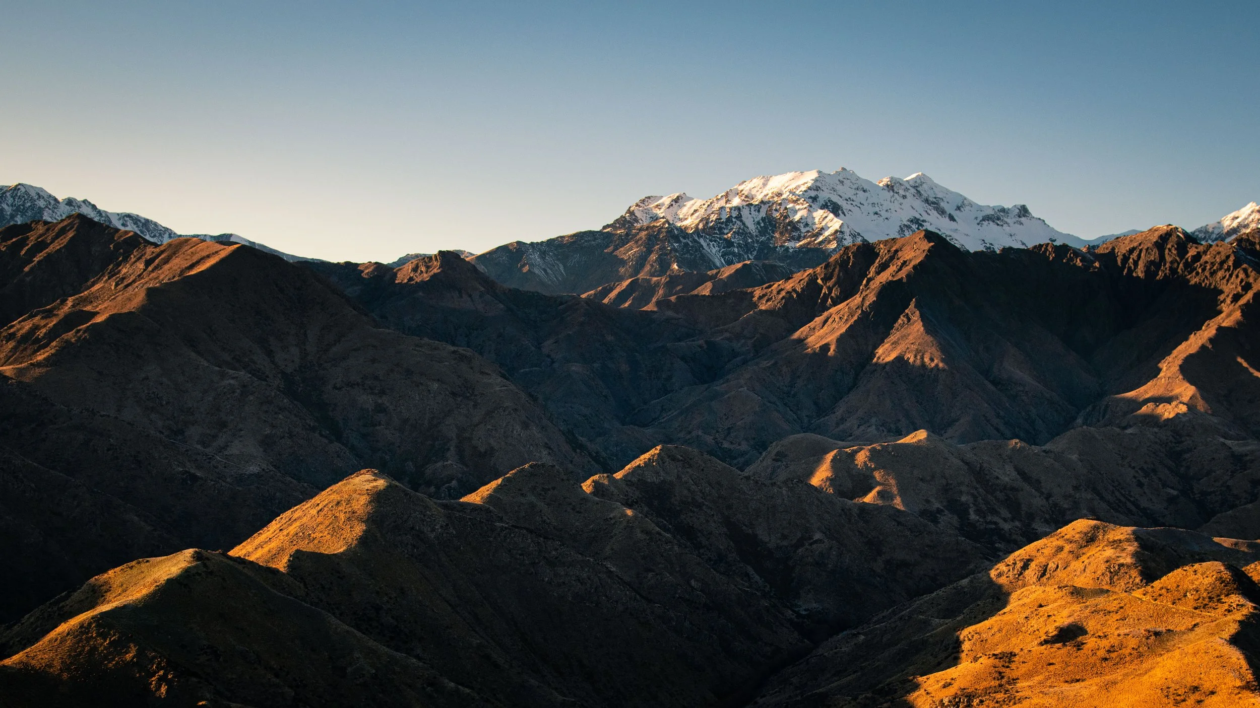 Mountain landscape with rugged brown and green hills in the foreground and snow-capped peaks in the background under a clear blue sky.