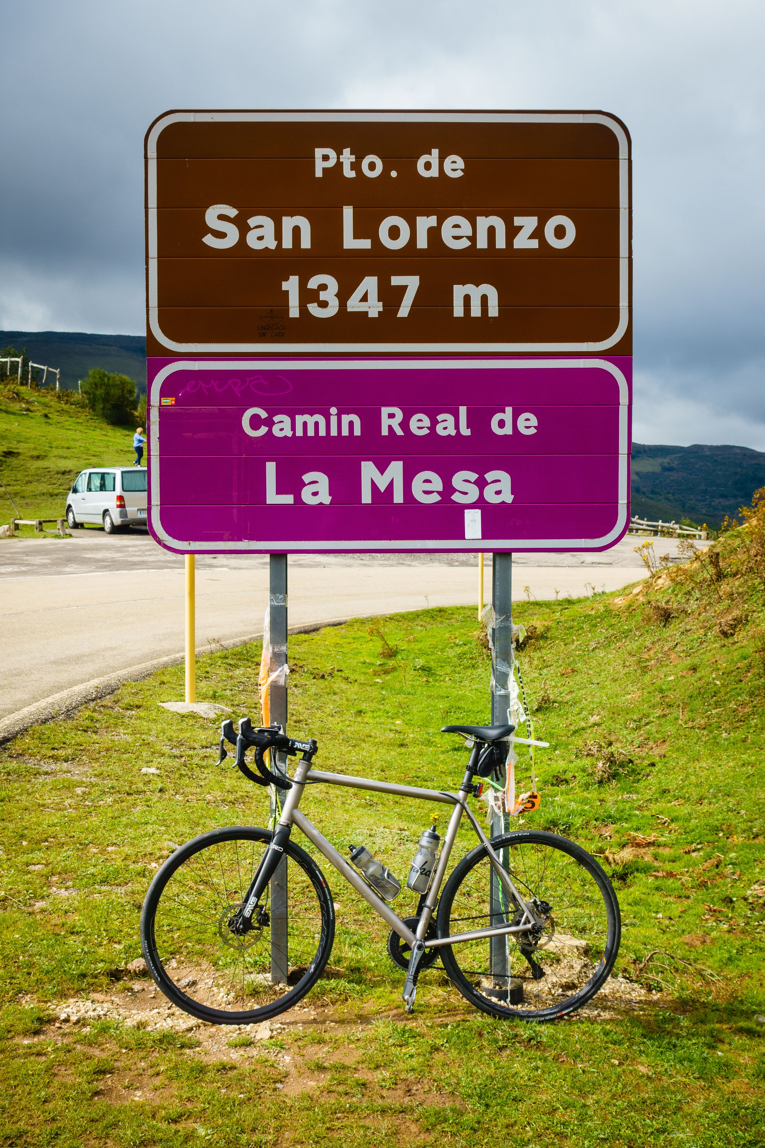 Road sign with brown top section reading 'Pto. de San Lorenzo 1347 m' and purple bottom section reading 'Camin Rea de La Mesa', with a bicycle parked in front on grass, in a mountainous landscape.