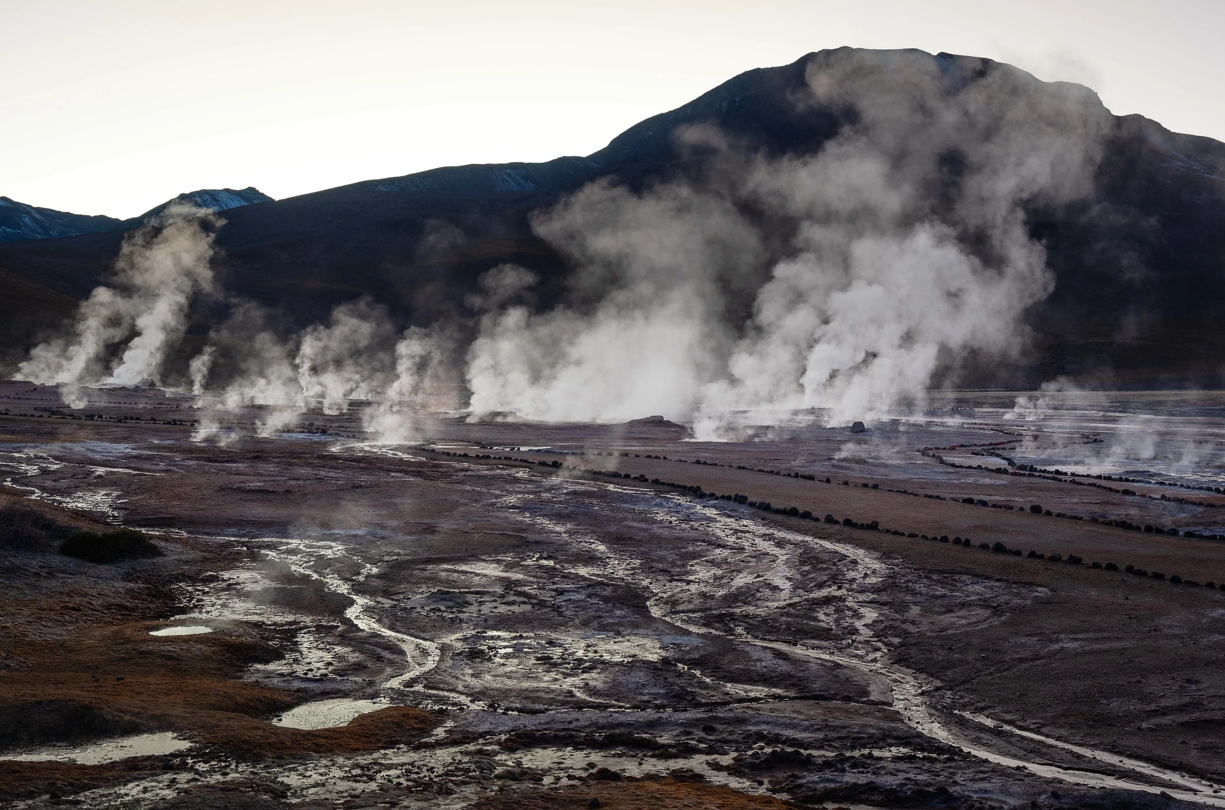 A fumarole field with steam rising from the ground in a valley, with mountains in the background.