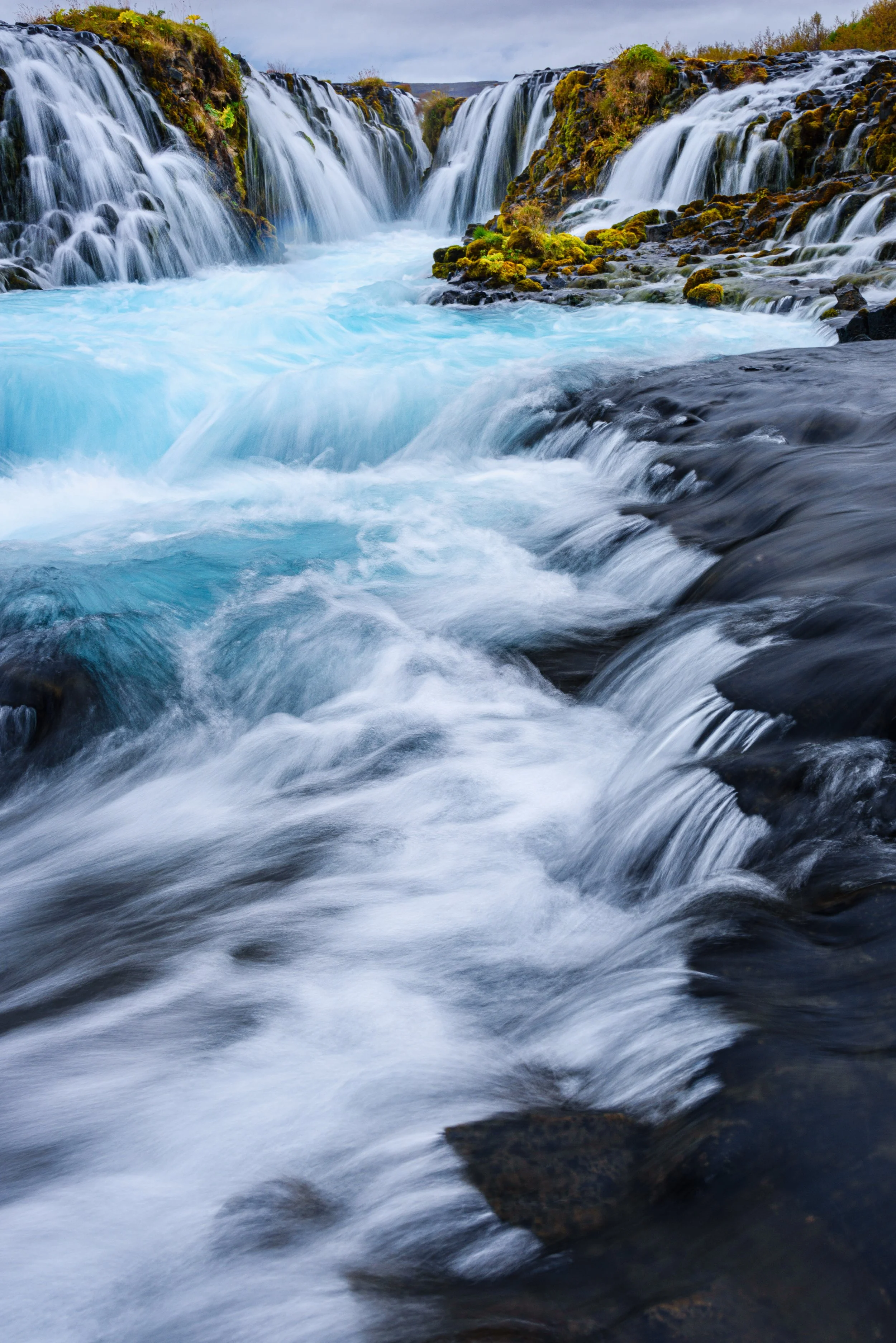 A waterfall with cascading water flowing over rocks, surrounded by moss and greenery, under a cloudy sky.